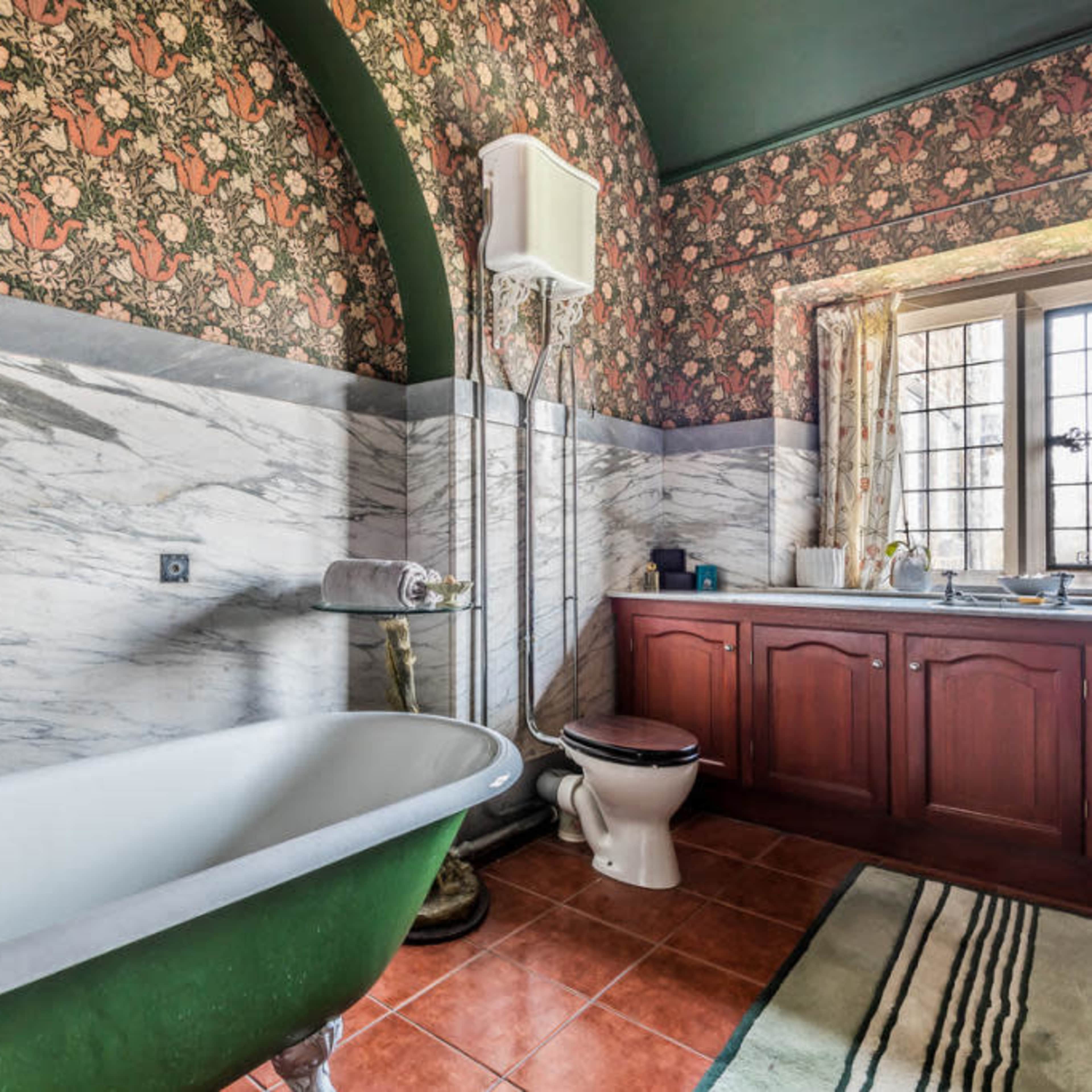 A vintage bathroom featuring a clawfoot bathtub, marble walls, a wooden vanity, and patterned wallpaper.