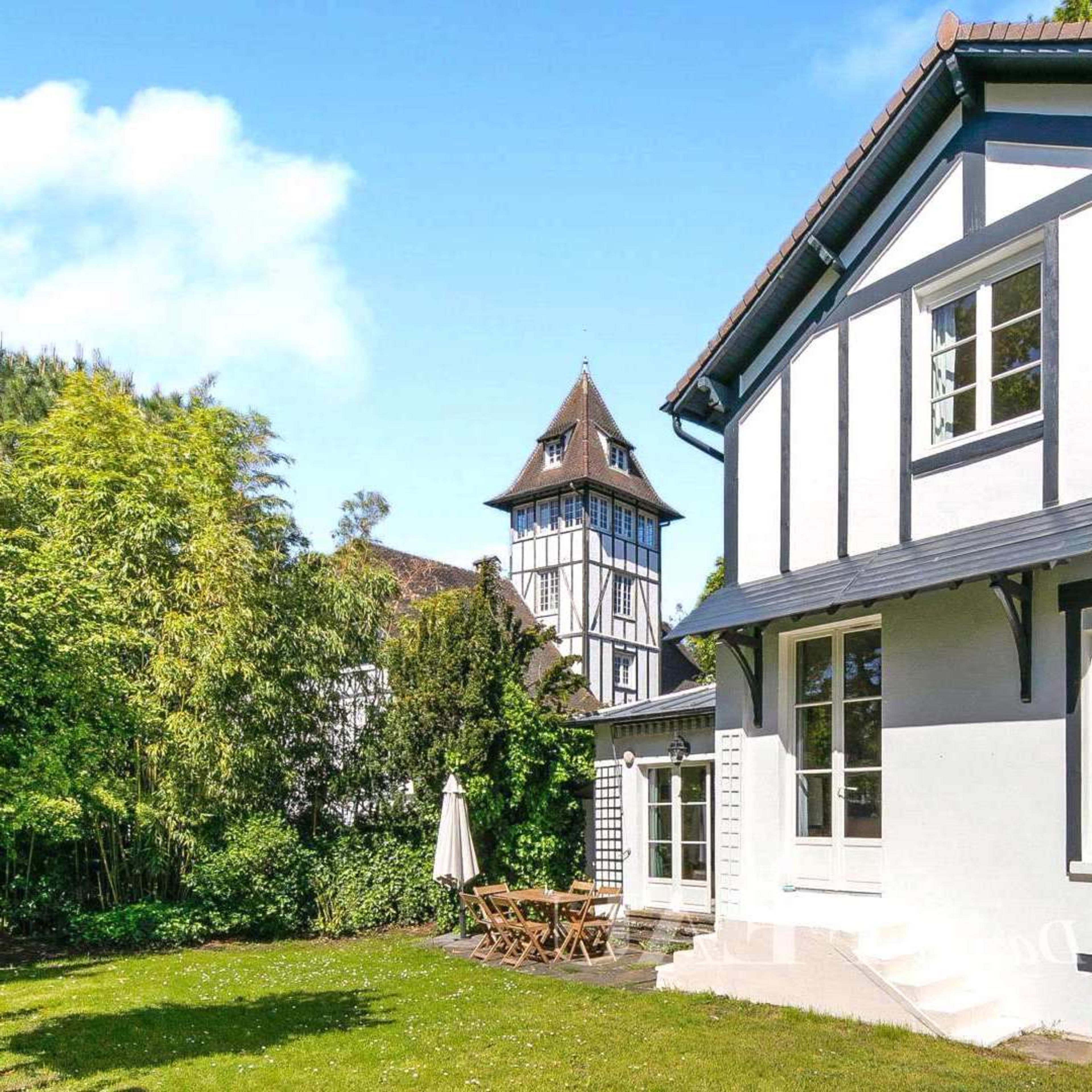 A two-story house with a white exterior and black accents, alongside a tall, pointed tower, set in a green garden with outdoor furniture.
