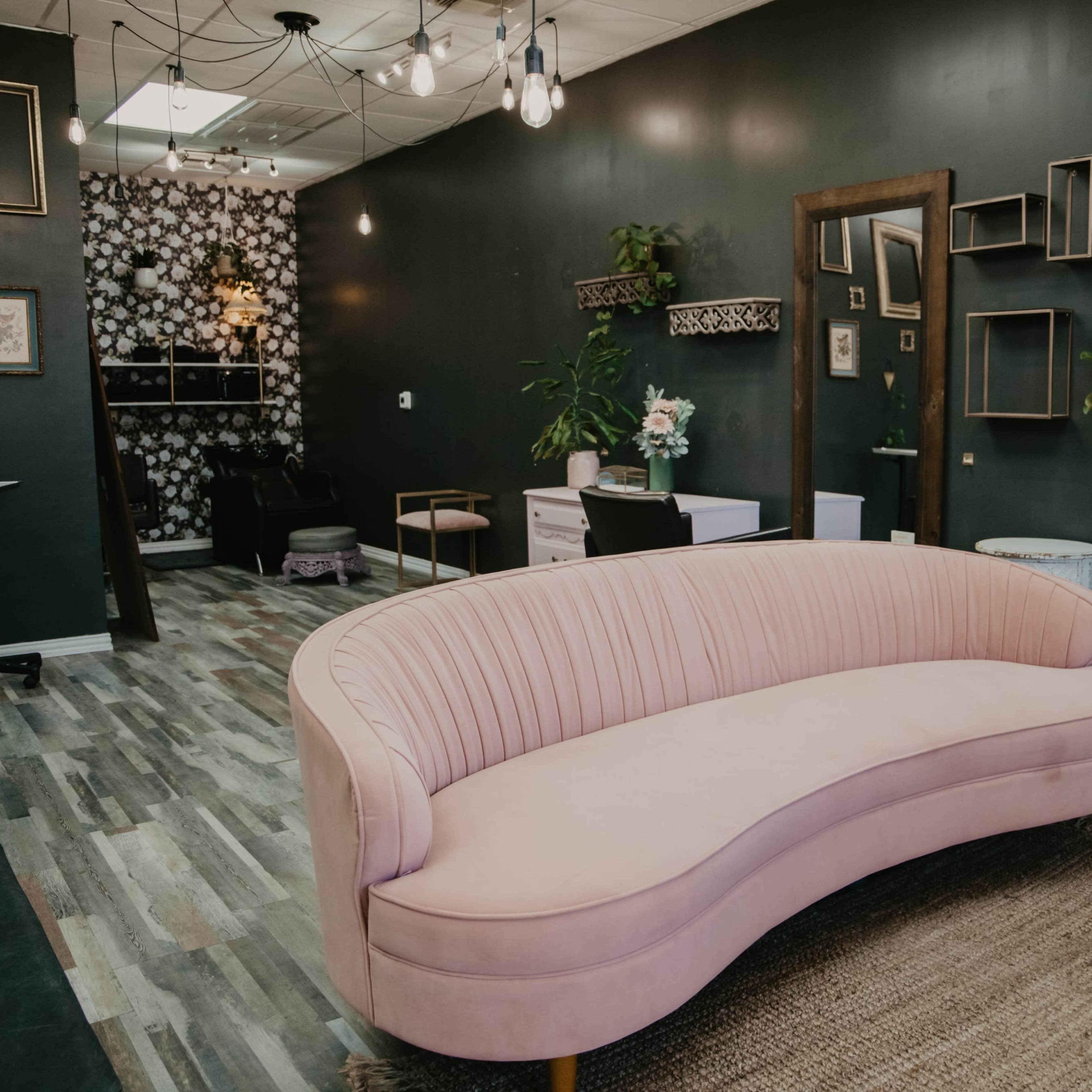 A stylish salon interior featuring a pink curved sofa, patterned wallpaper, and shelves filled with beauty products.