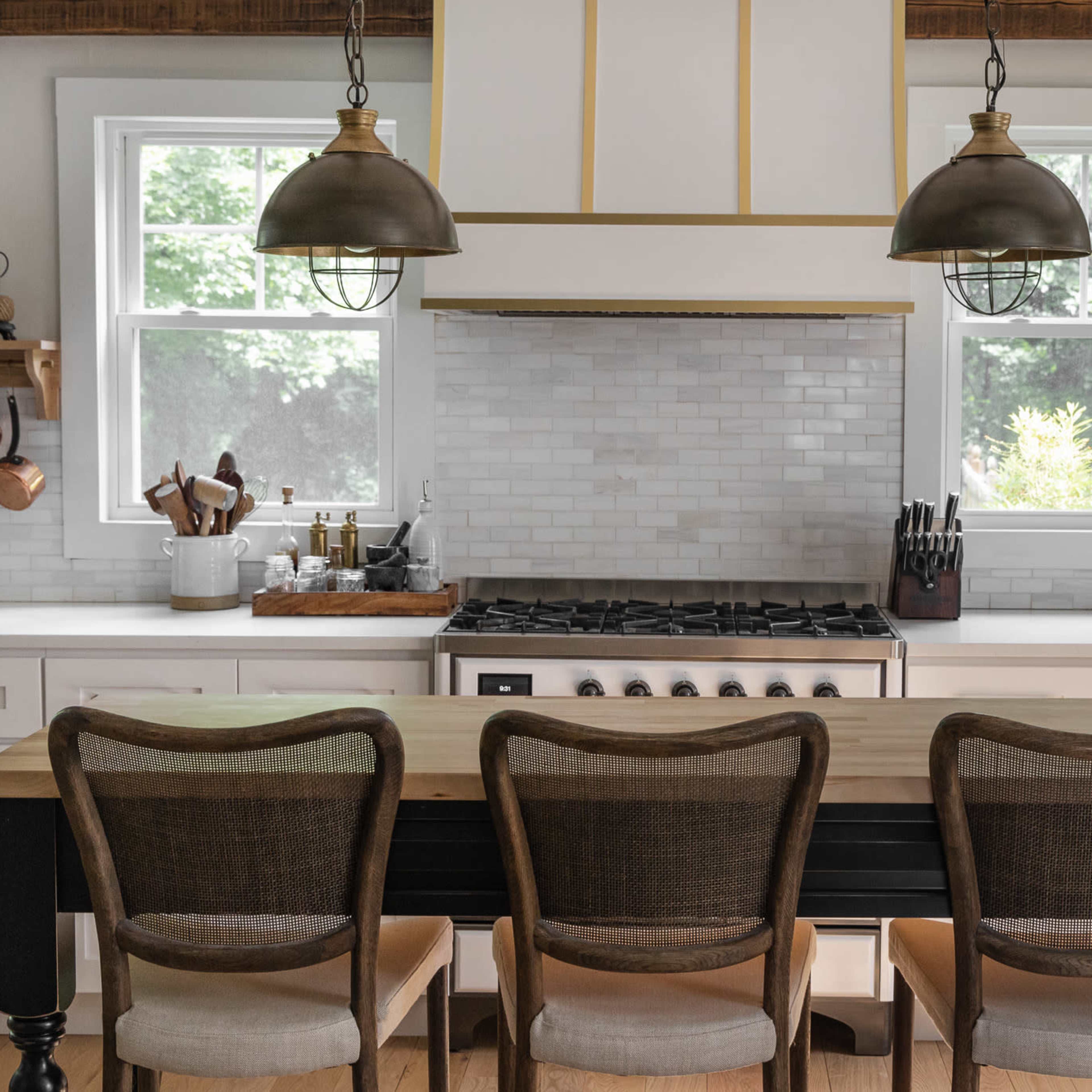 A modern kitchen features a central island with three seated wooden chairs, white cabinetry, a stainless steel stove, and pendant lighting above.
