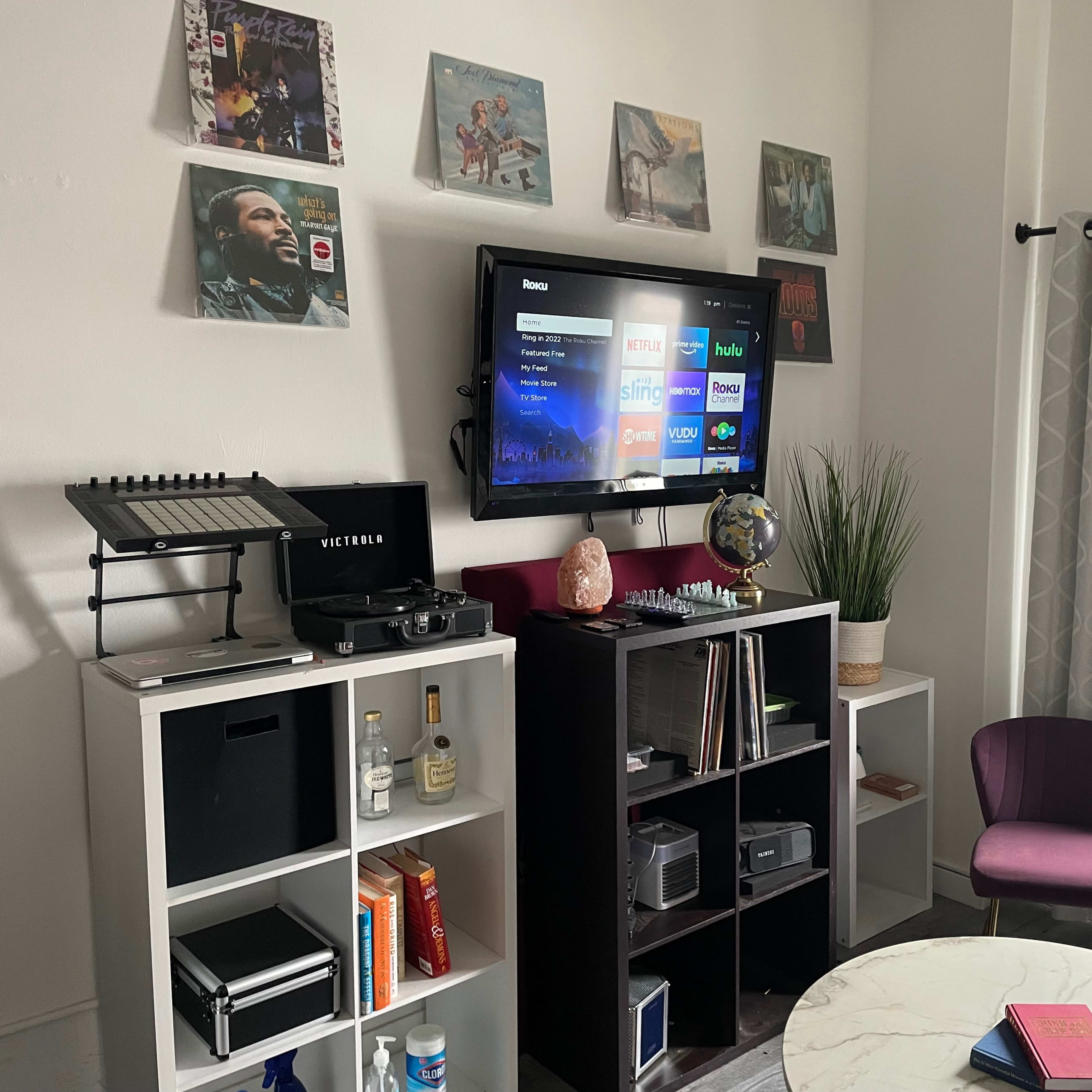 A living room corner with a television mounted on the wall above a media console, surrounded by vinyl records and decorative items, and featuring a small seating area with a round table and a purple chair.
