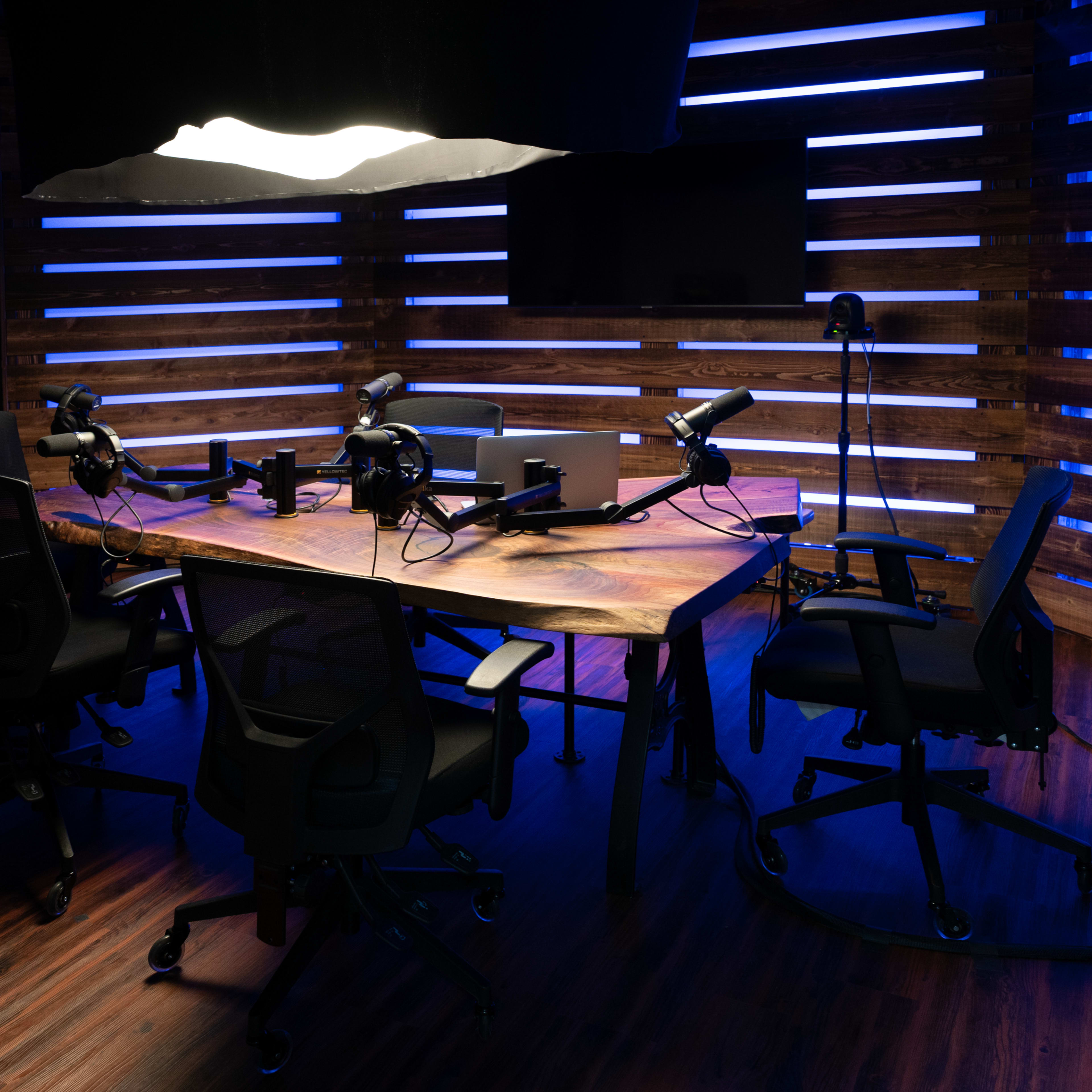 A podcast recording studio features a large wooden table surrounded by four black chairs, with microphones and equipment set up in a dimly lit space accentuated by blue lighting.