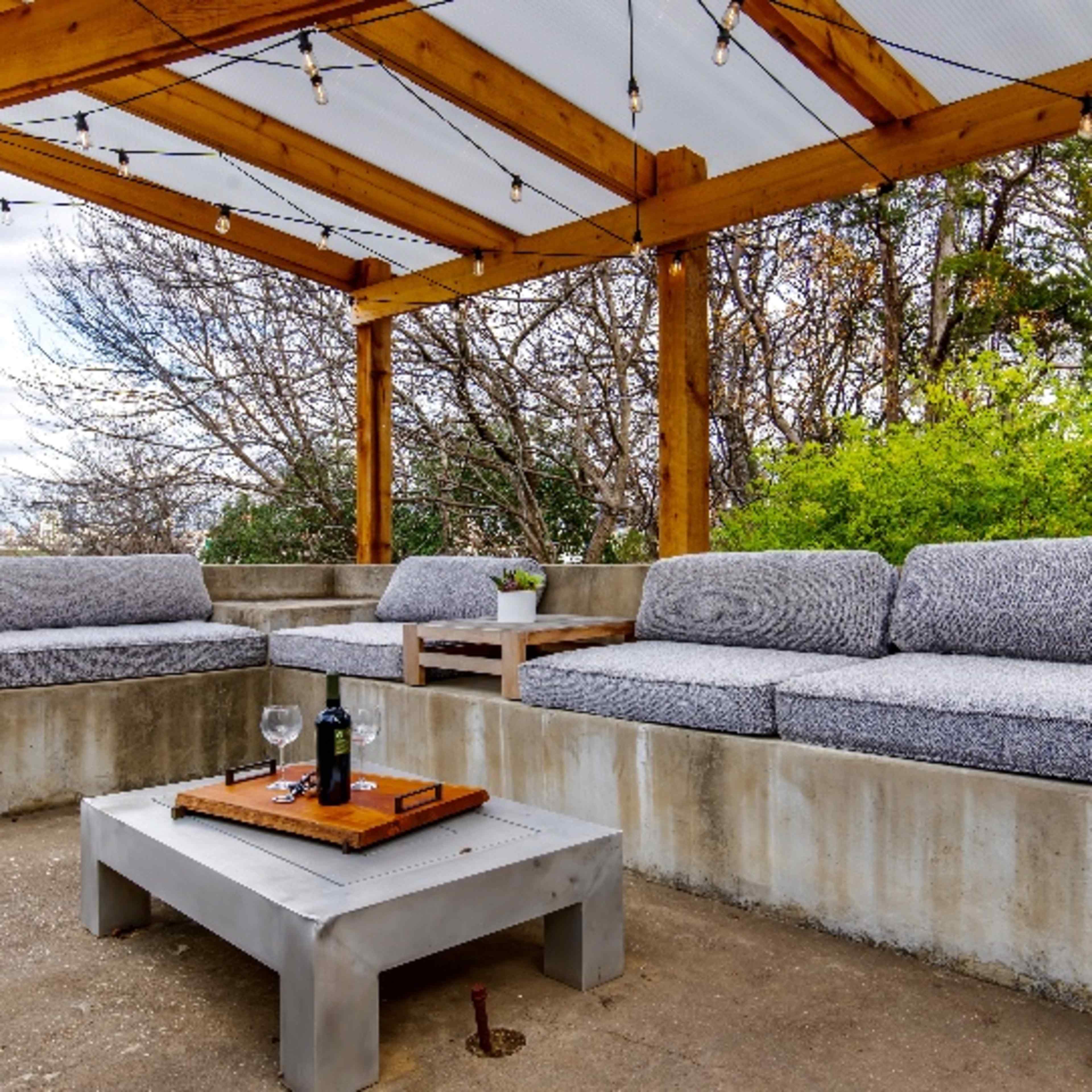 A shaded outdoor seating area with a concrete sofa, wooden beams, and a coffee table holding a wine bottle and glass.