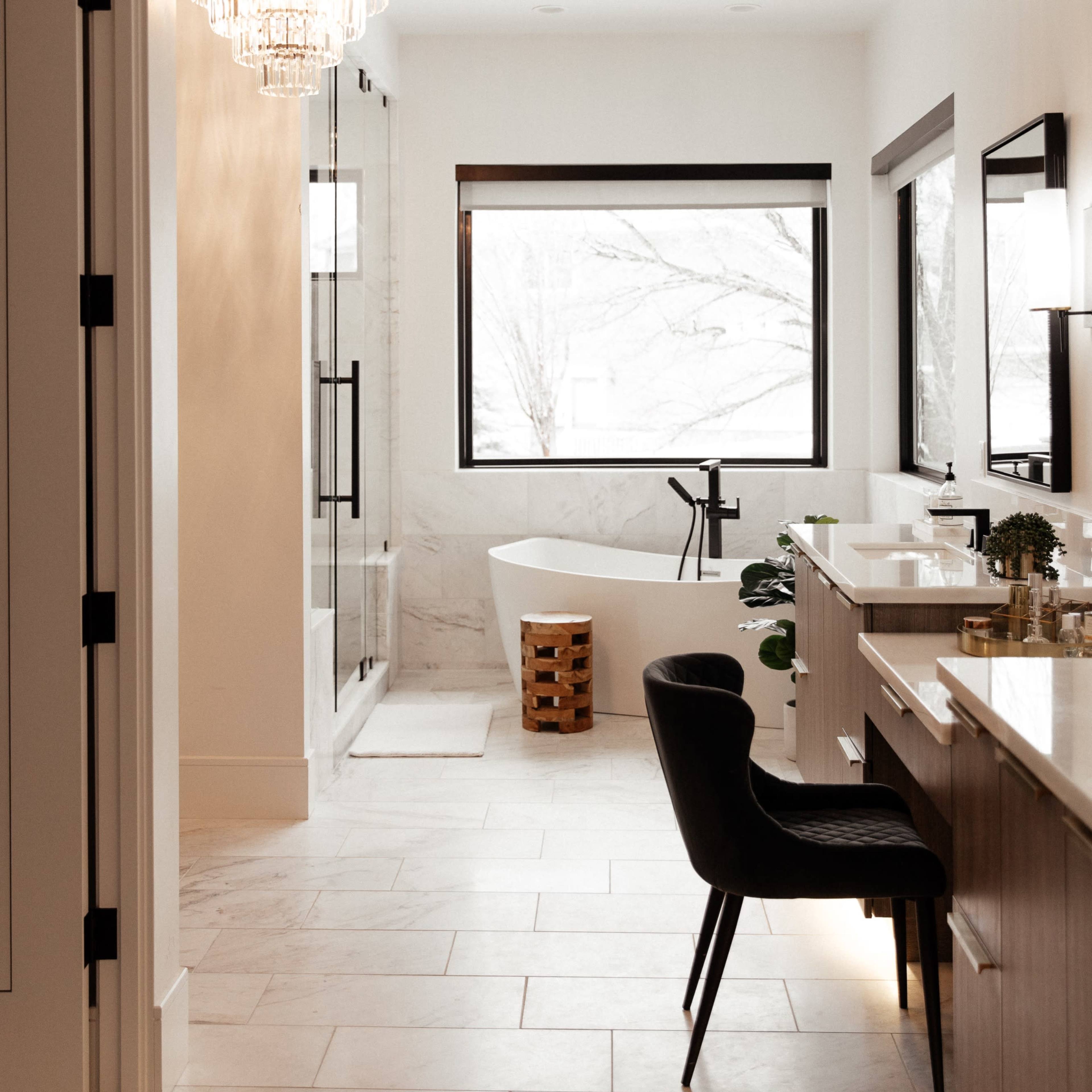 A modern bathroom features a freestanding tub, a glass-enclosed shower, and a chandelier, with ample natural light coming through large windows.