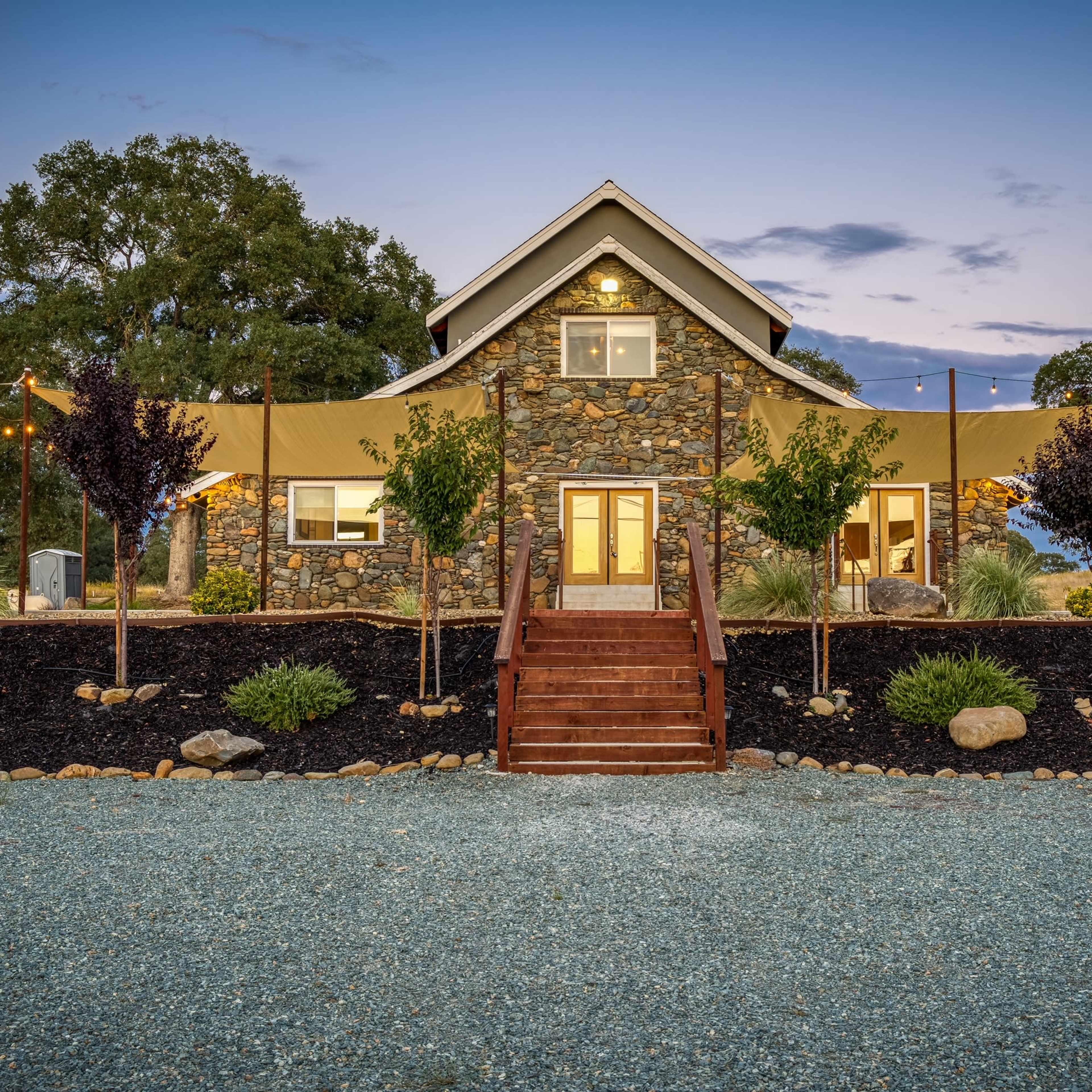 A stone house with a staircase and outdoor canopy is set against a twilight sky, surrounded by landscaped plants and gravel pathways.