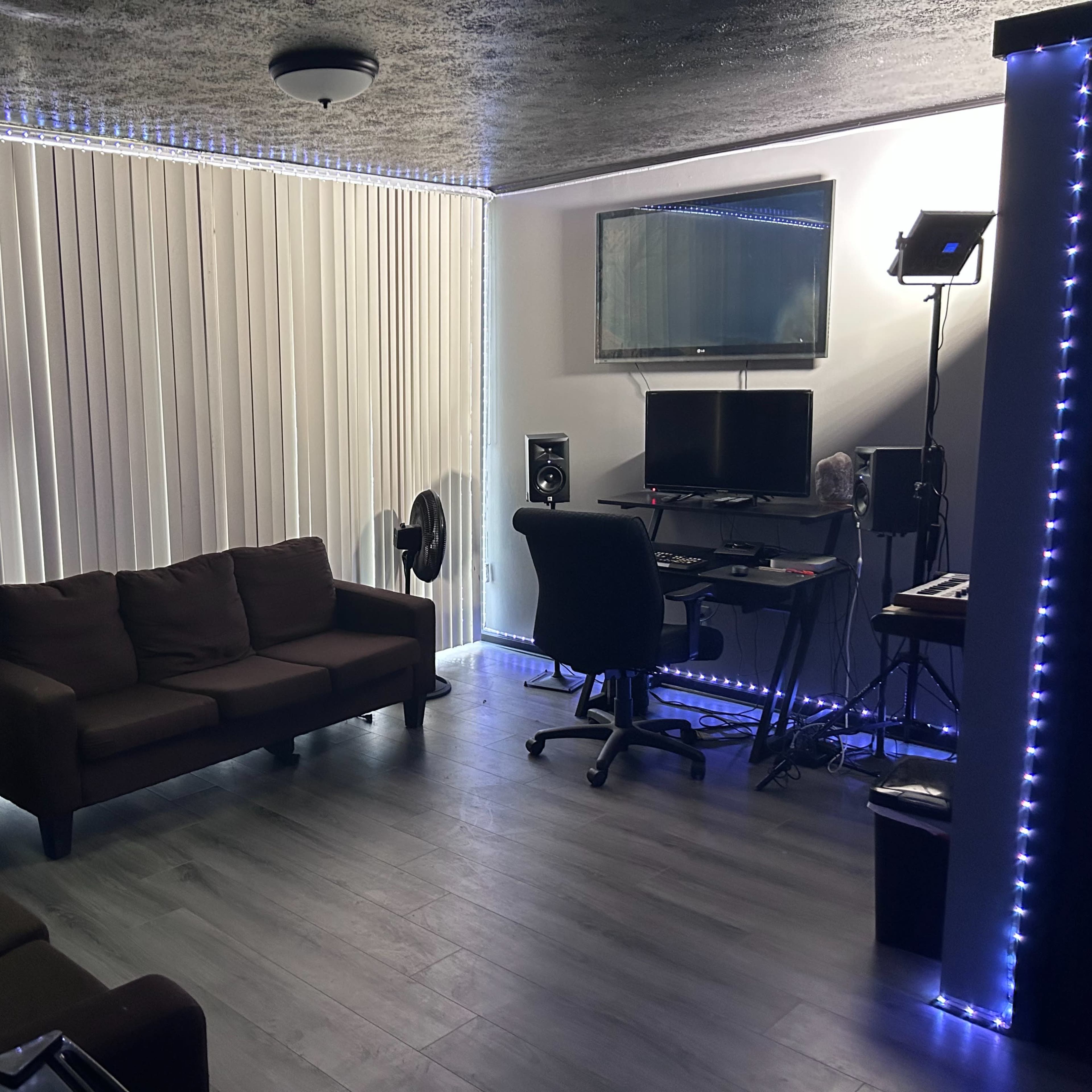 A small room features a brown couch, a desk with a computer and music equipment, a fan, and a tall mirrored cabinet, all illuminated by LED strip lights.