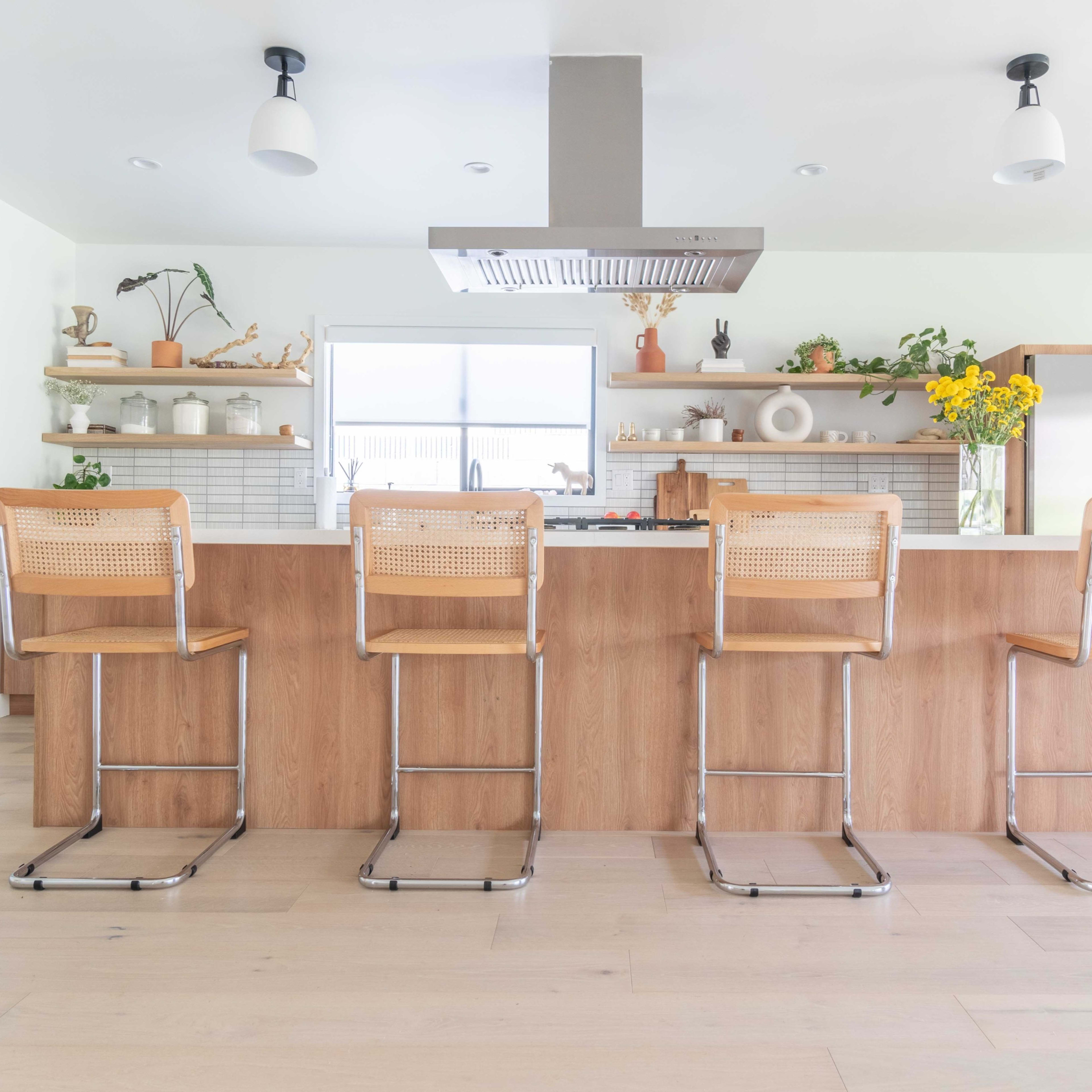 A modern kitchen with a long wooden island featuring four metal and wood bar stools, surrounded by light-colored walls and various decorative items on shelves.