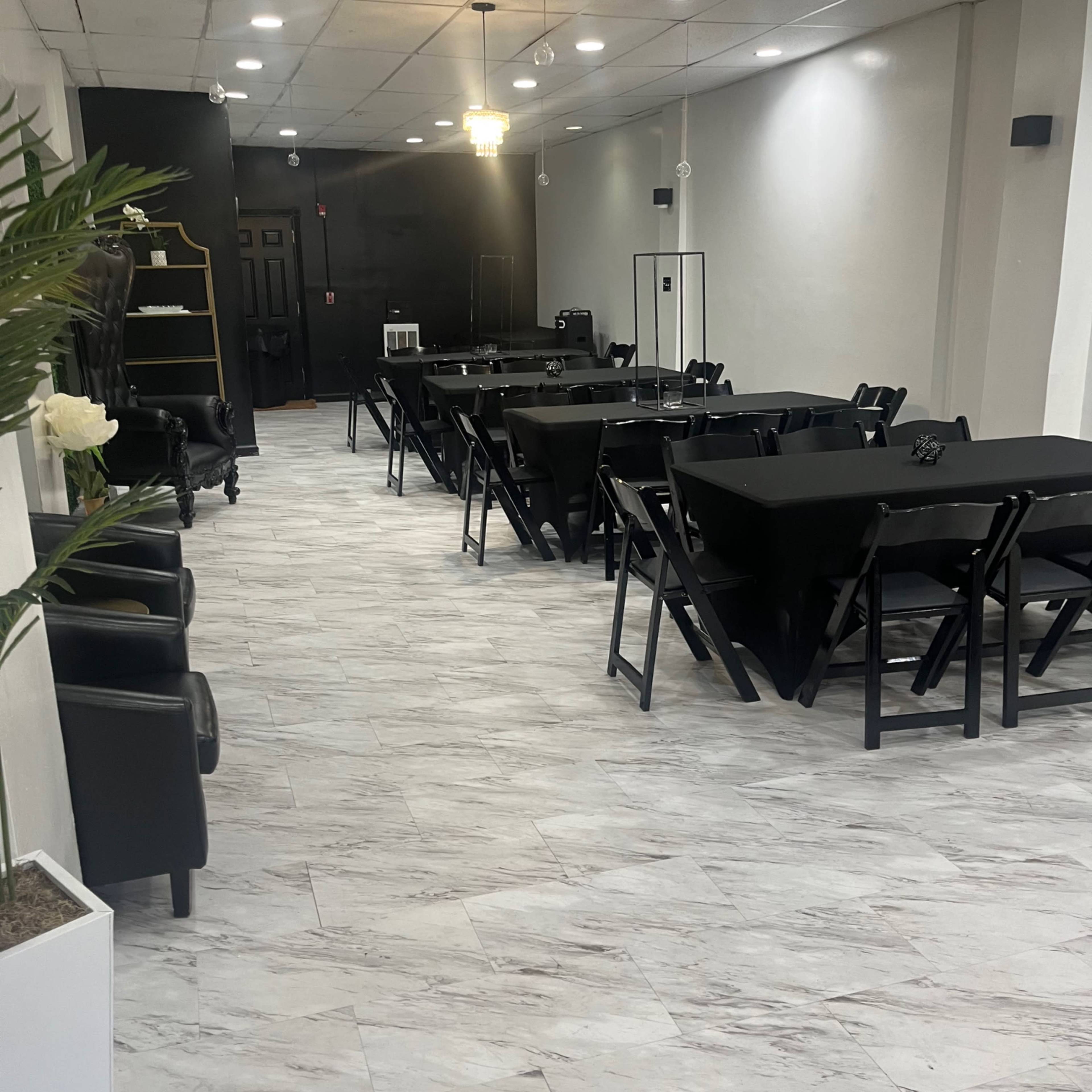 A spacious event room with several black folding tables and chairs arranged in rows, featuring light-colored tiles and a black accent wall.