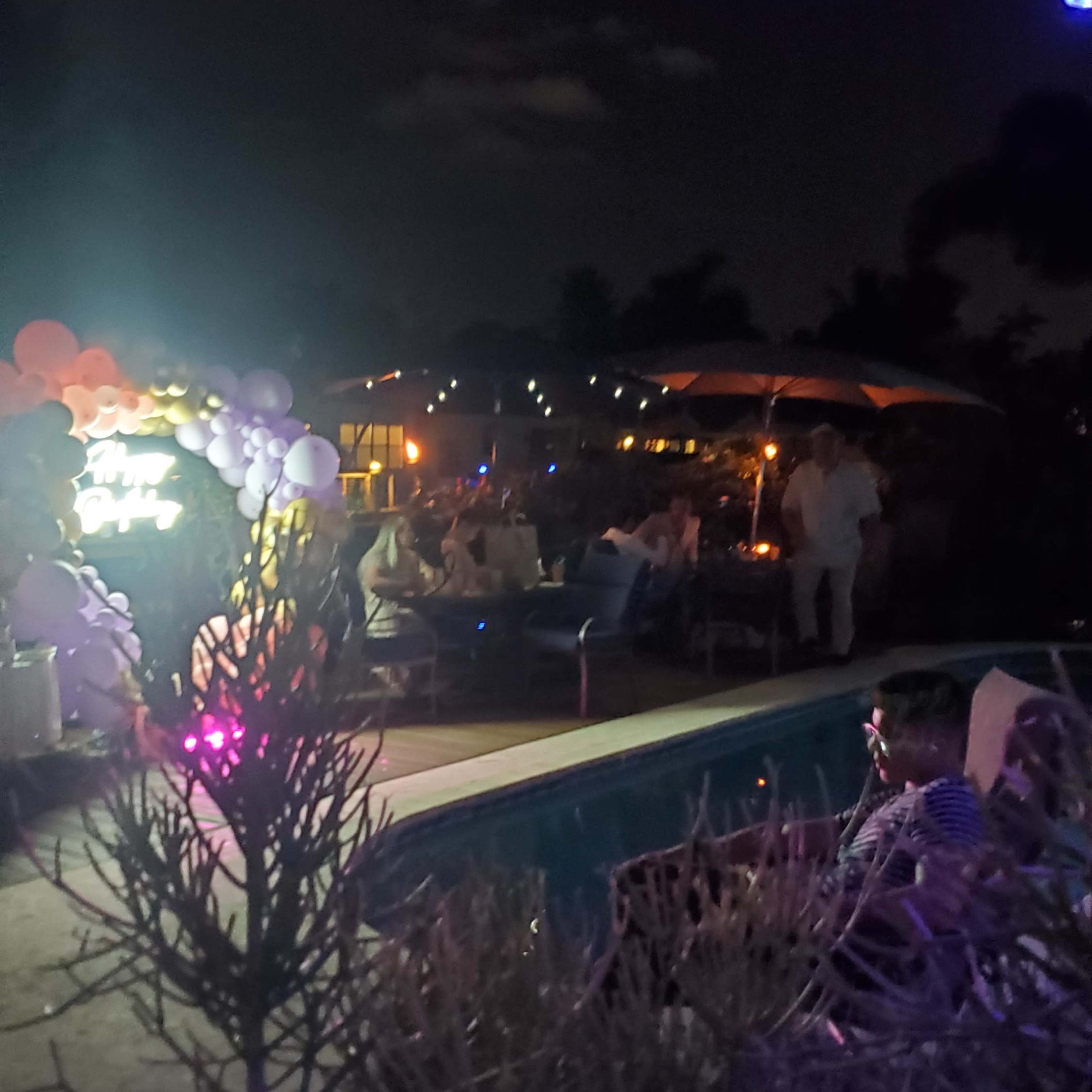 A nighttime outdoor party scene with tables, balloons, and dim lighting around a swimming pool.