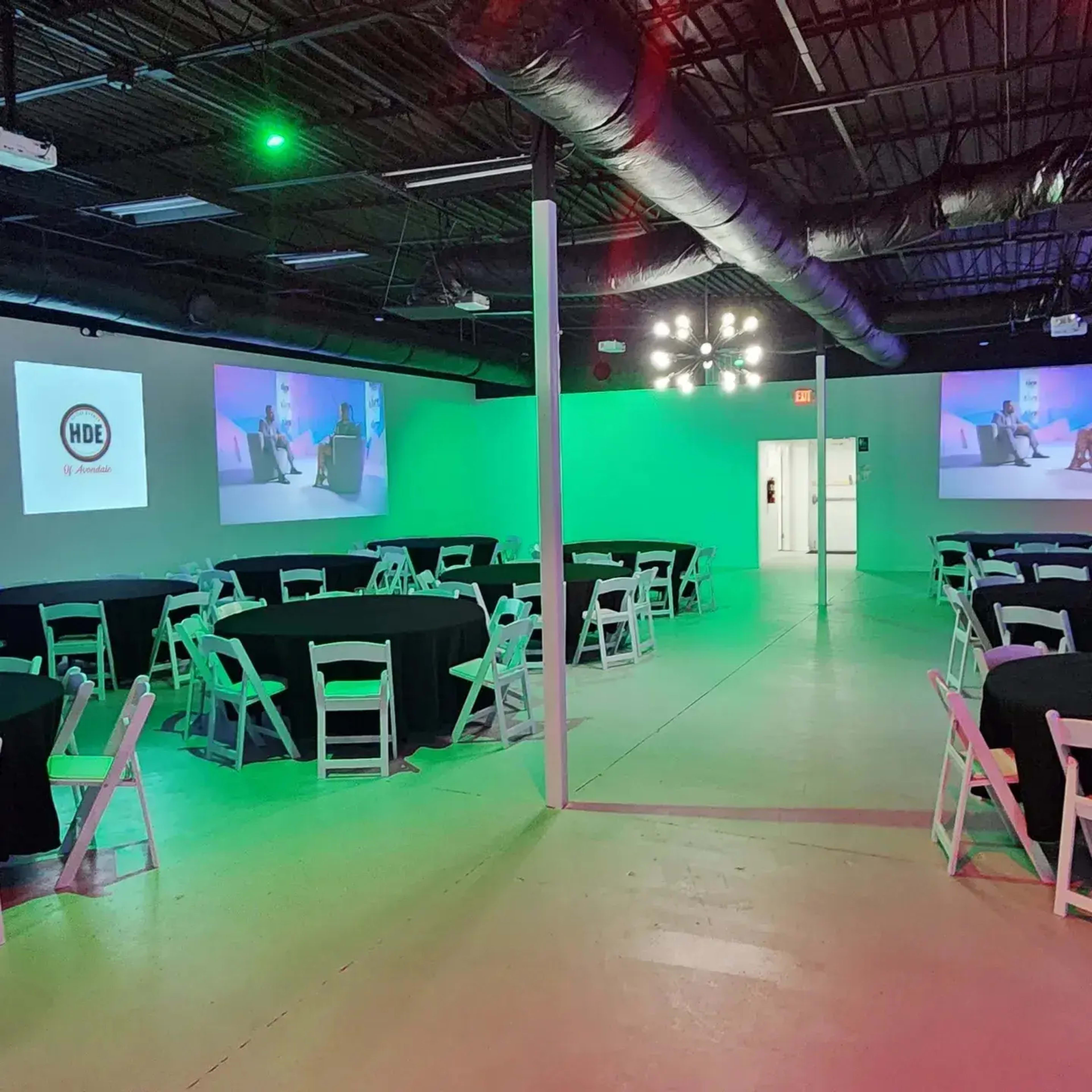 A spacious event room set up with multiple black round tables and white chairs, illuminated by green lights, with presentations displayed on two screens.