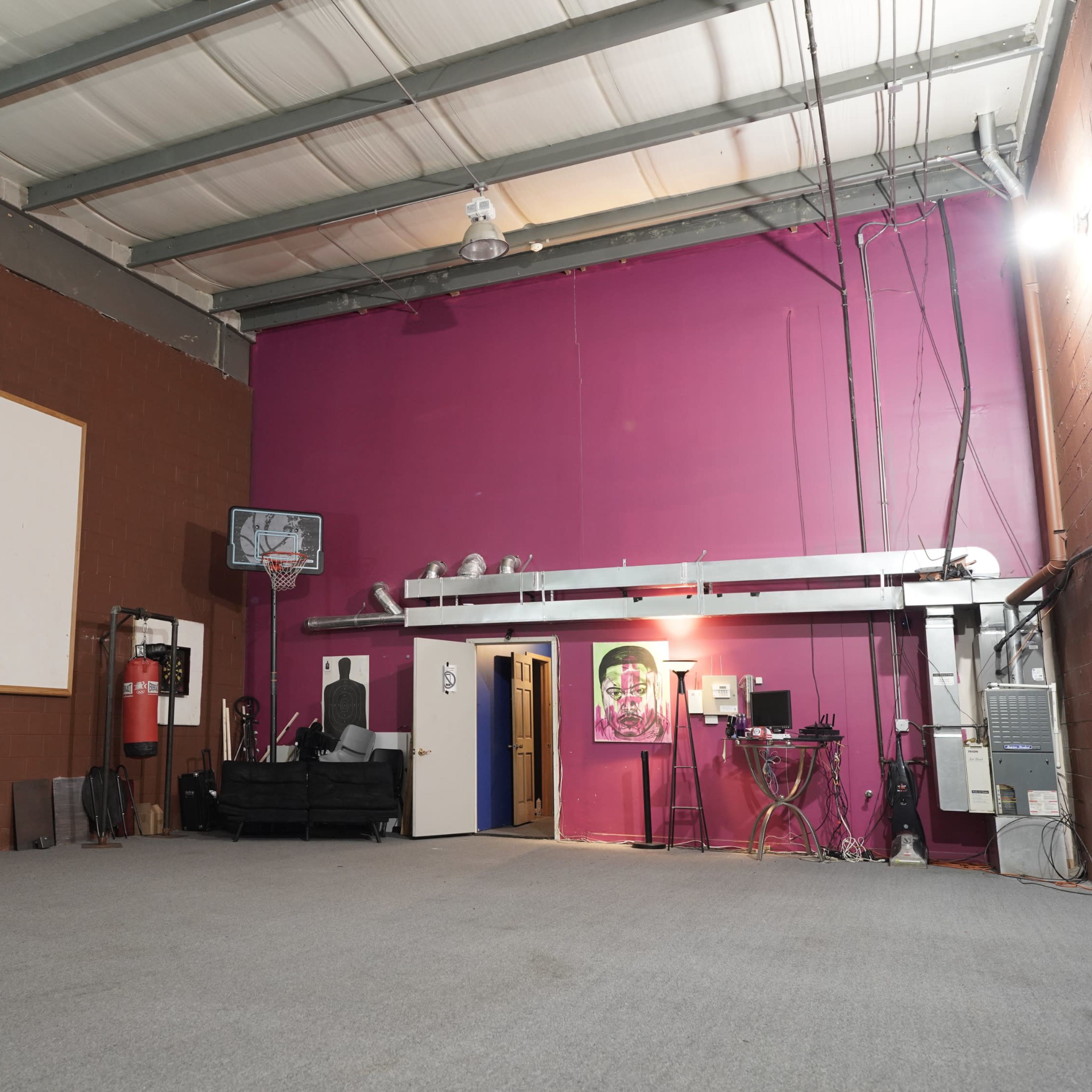 An empty room with a high ceiling, a large blank wall on the left, and a vibrant pink wall on the right, featuring a door and various equipment along the back wall.
