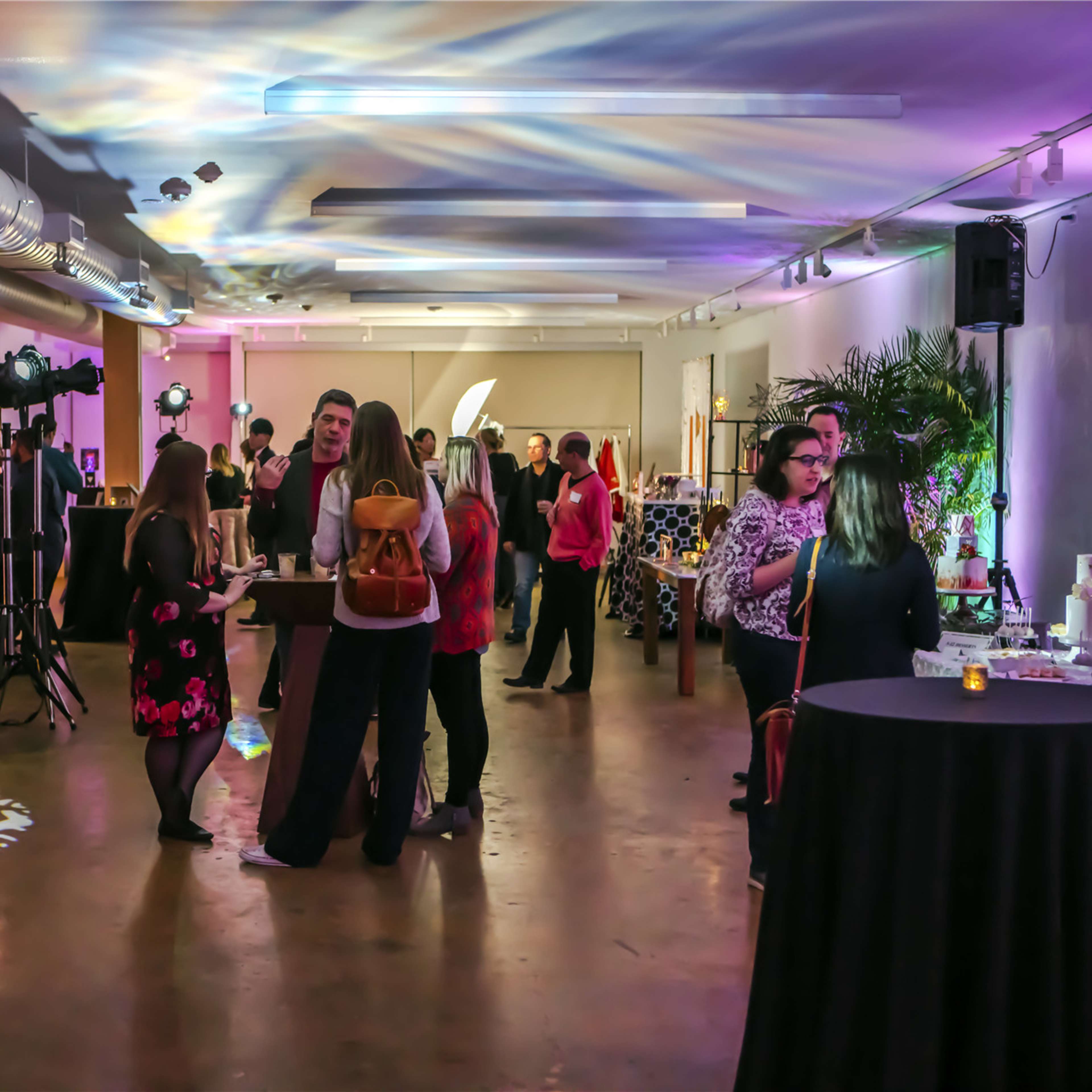 A spacious event venue filled with people mingling around cocktail tables, with colorful lighting and decorative elements creating an inviting atmosphere.
