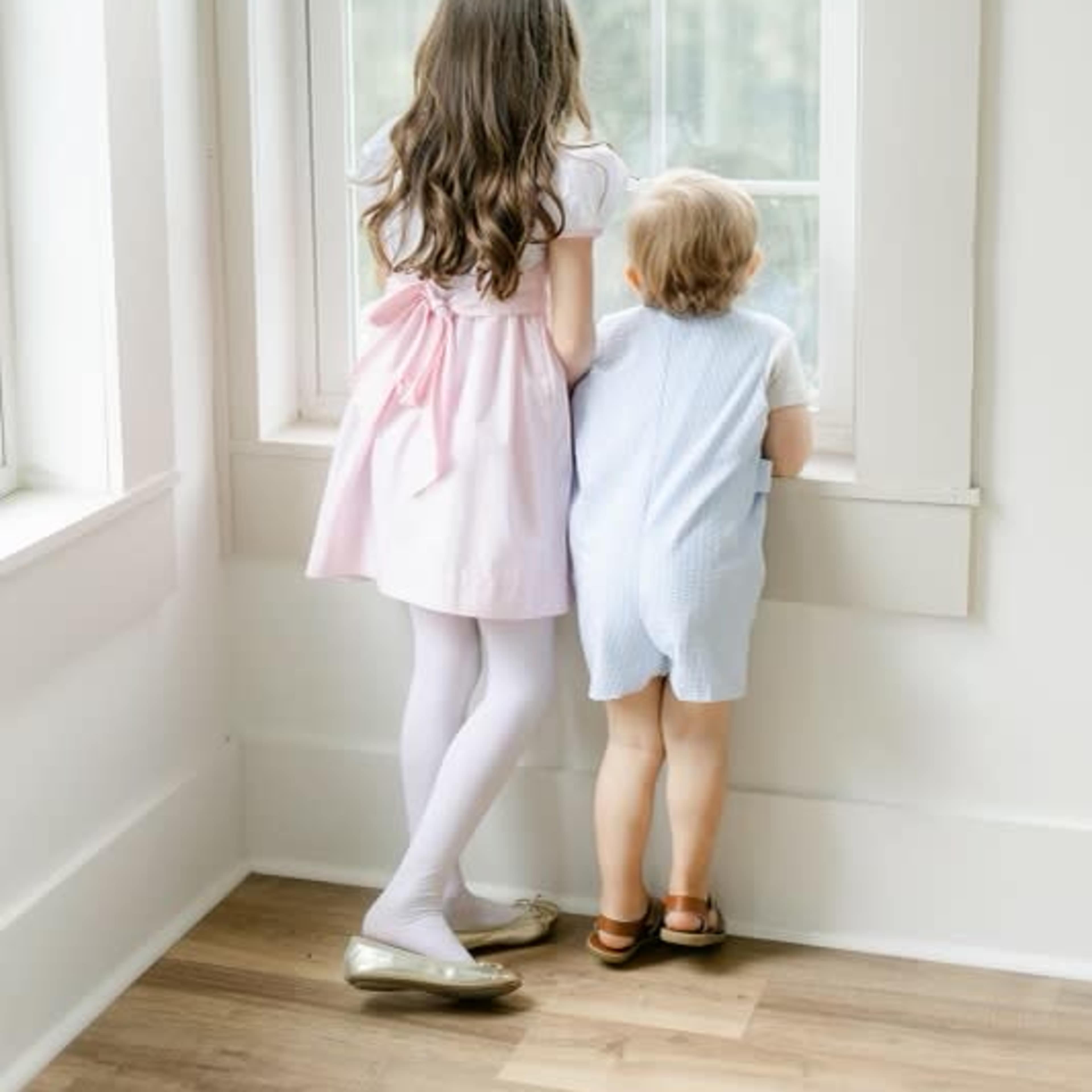 Two children stand by a large window, looking outside, with one wearing a pink dress and the other in a light blue outfit.