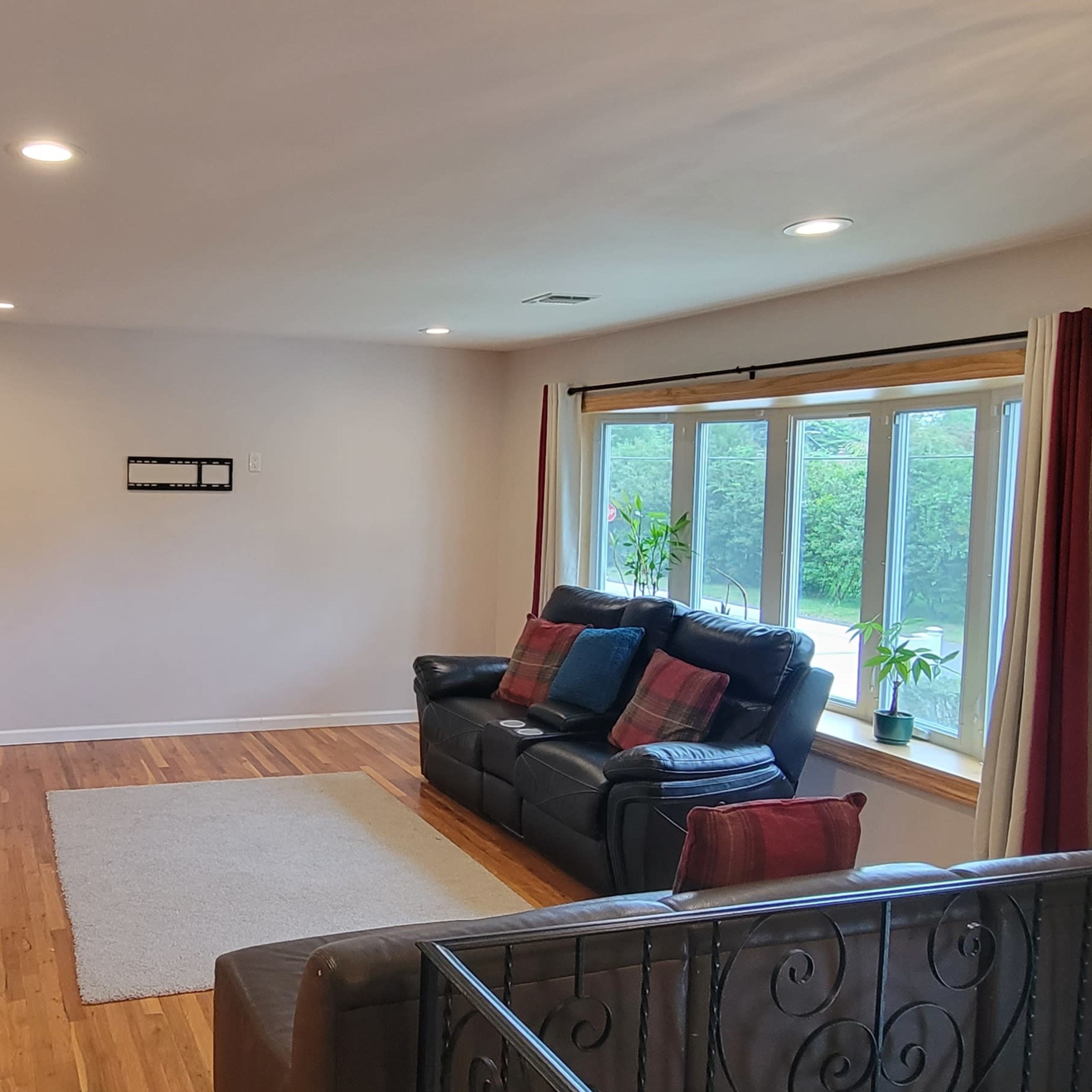A spacious living room with two black sofas, a light-colored rug on hardwood flooring, a table against the wall, and large windows allowing natural light to enter.