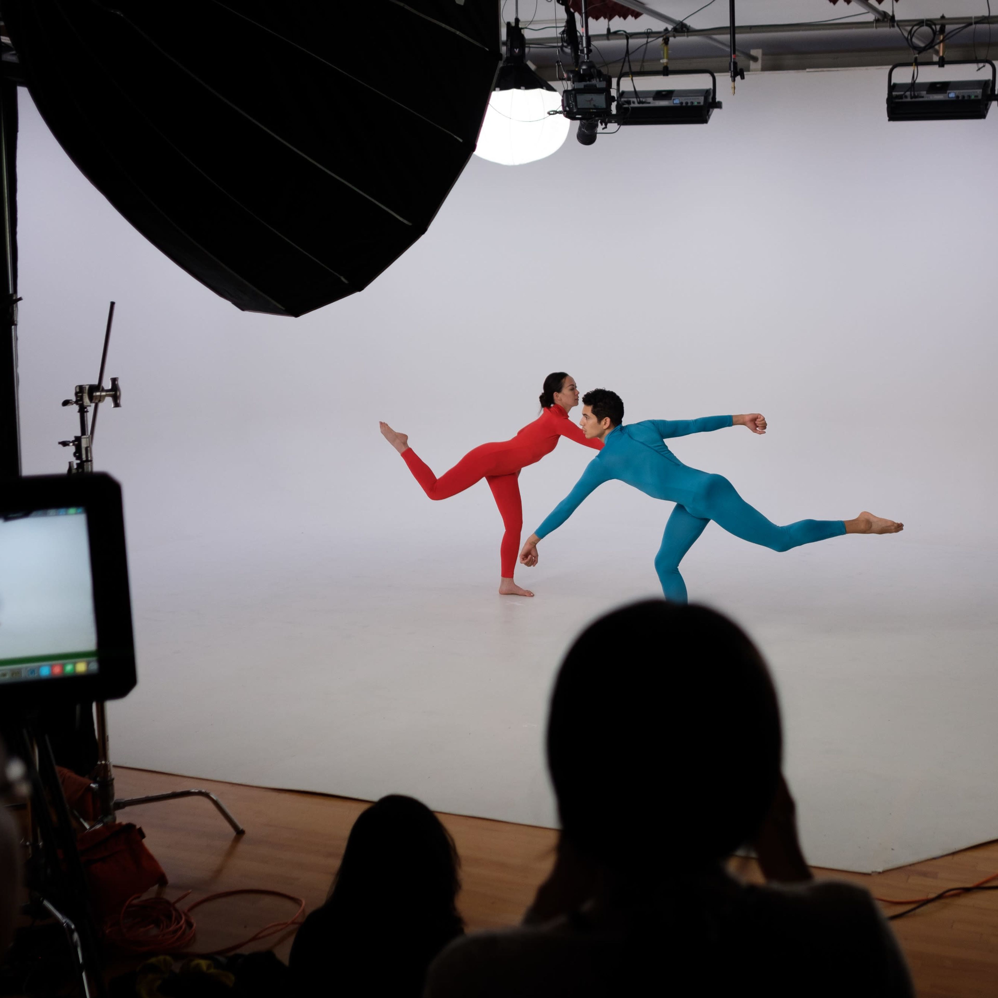 Two dancers in red and blue costumes perform a ballet pose in a photo studio with a plain white backdrop and camera equipment in the foreground.