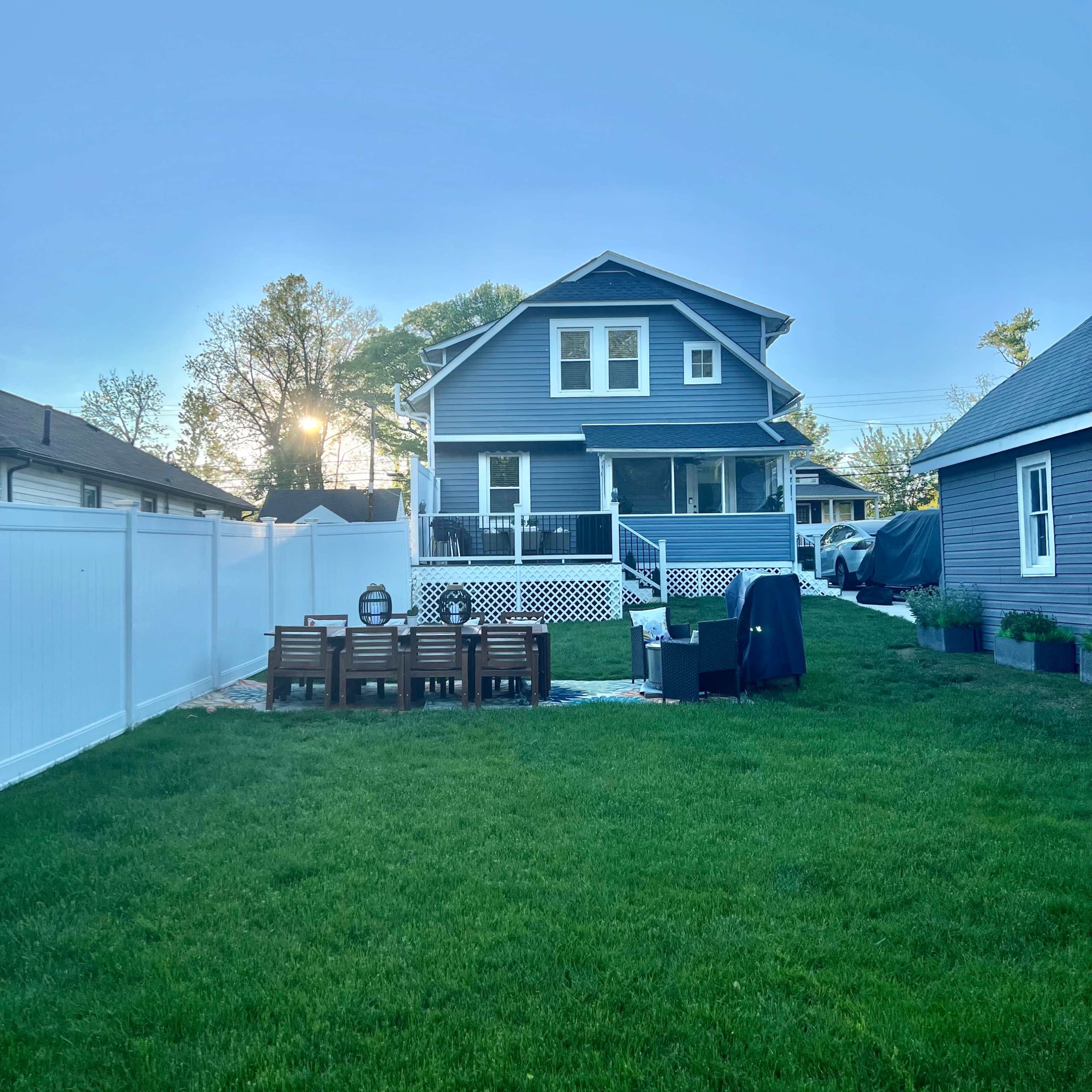 A blue two-story house with a porch overlooks a green lawn enclosed by a white fence, featuring a patio set in the foreground.