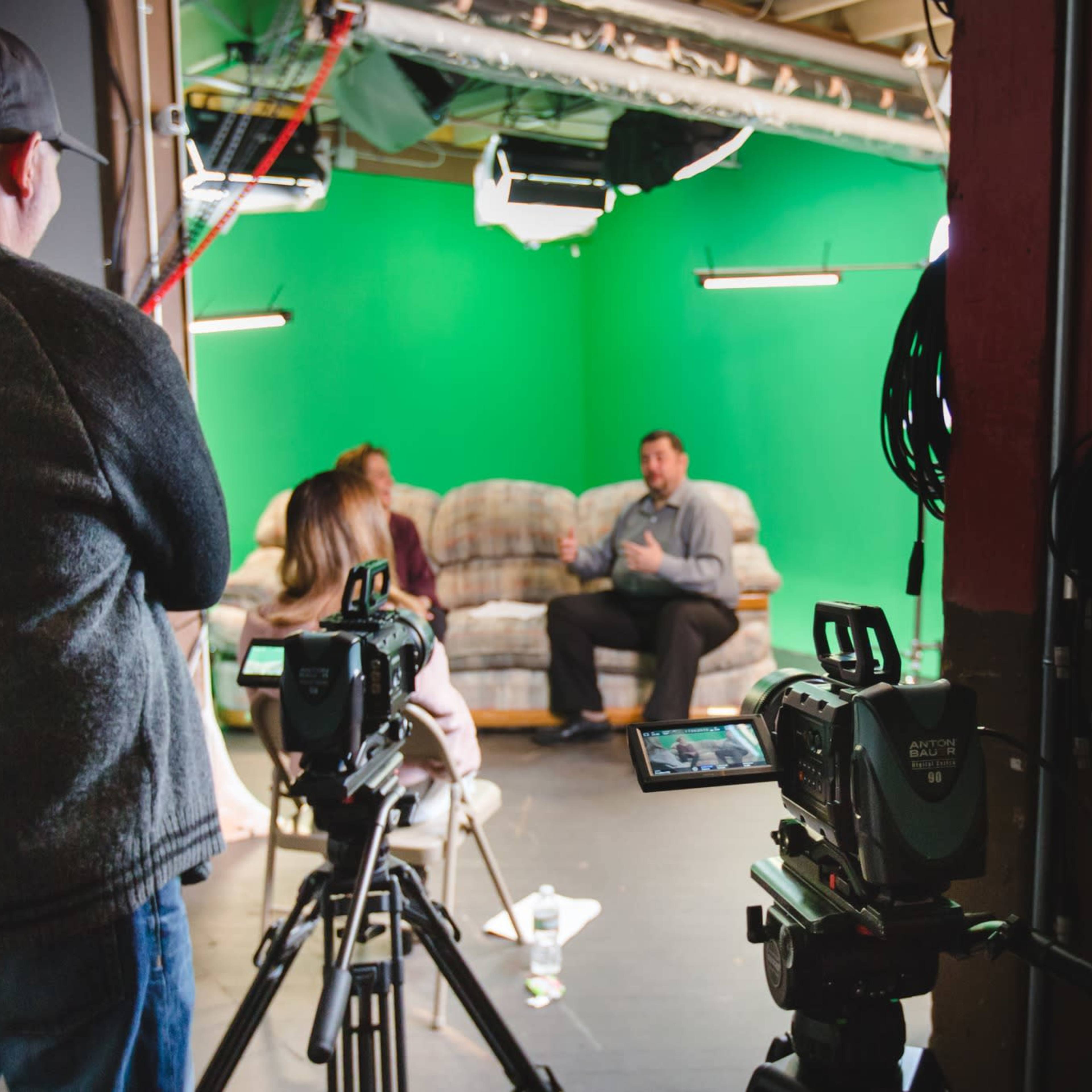 A man in a sweater operates a camera while a woman and a man are seated on a couch in front of a green screen during a video shoot.