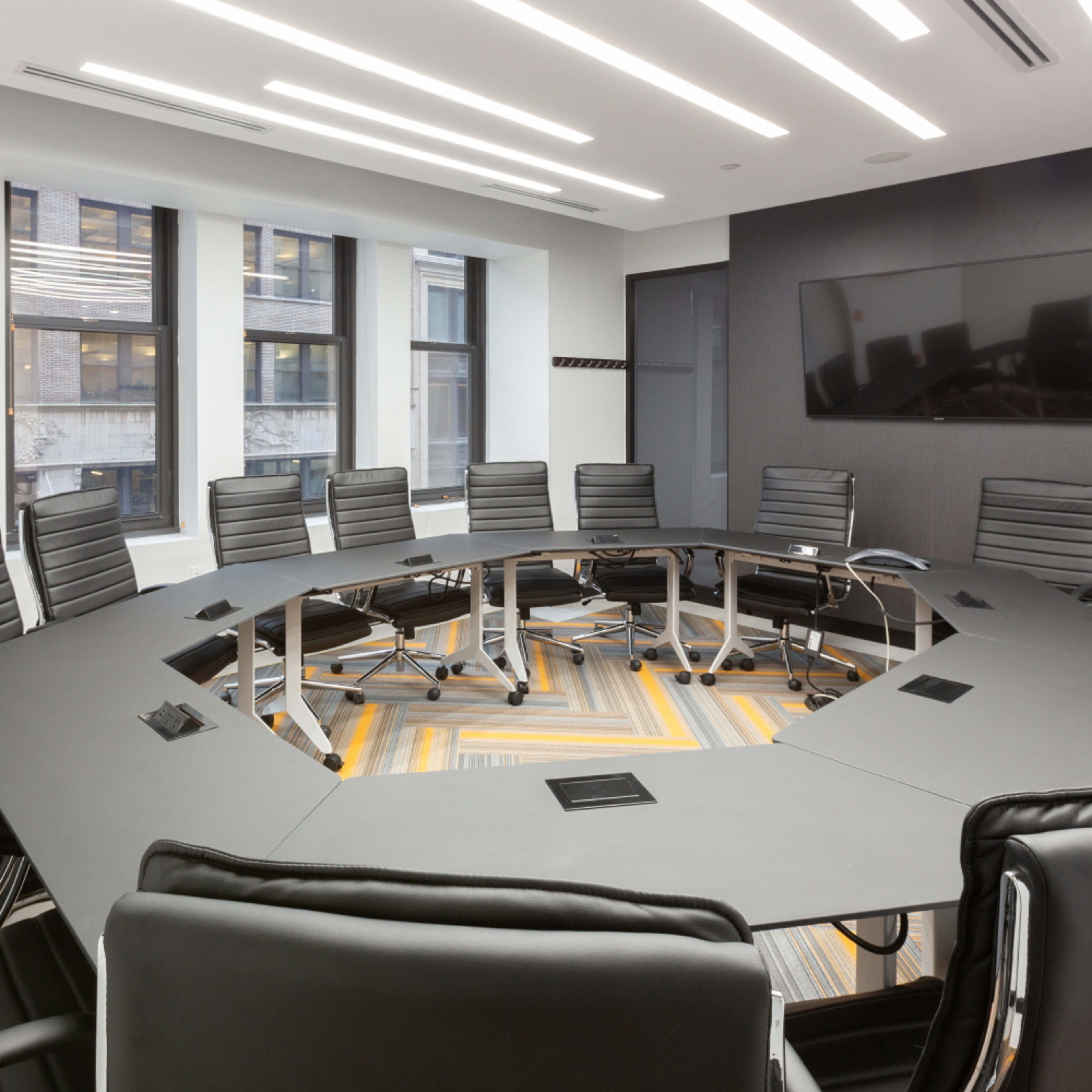 A large, round meeting table surrounded by black chairs is shown in a modern conference room with large windows and a wall-mounted screen.