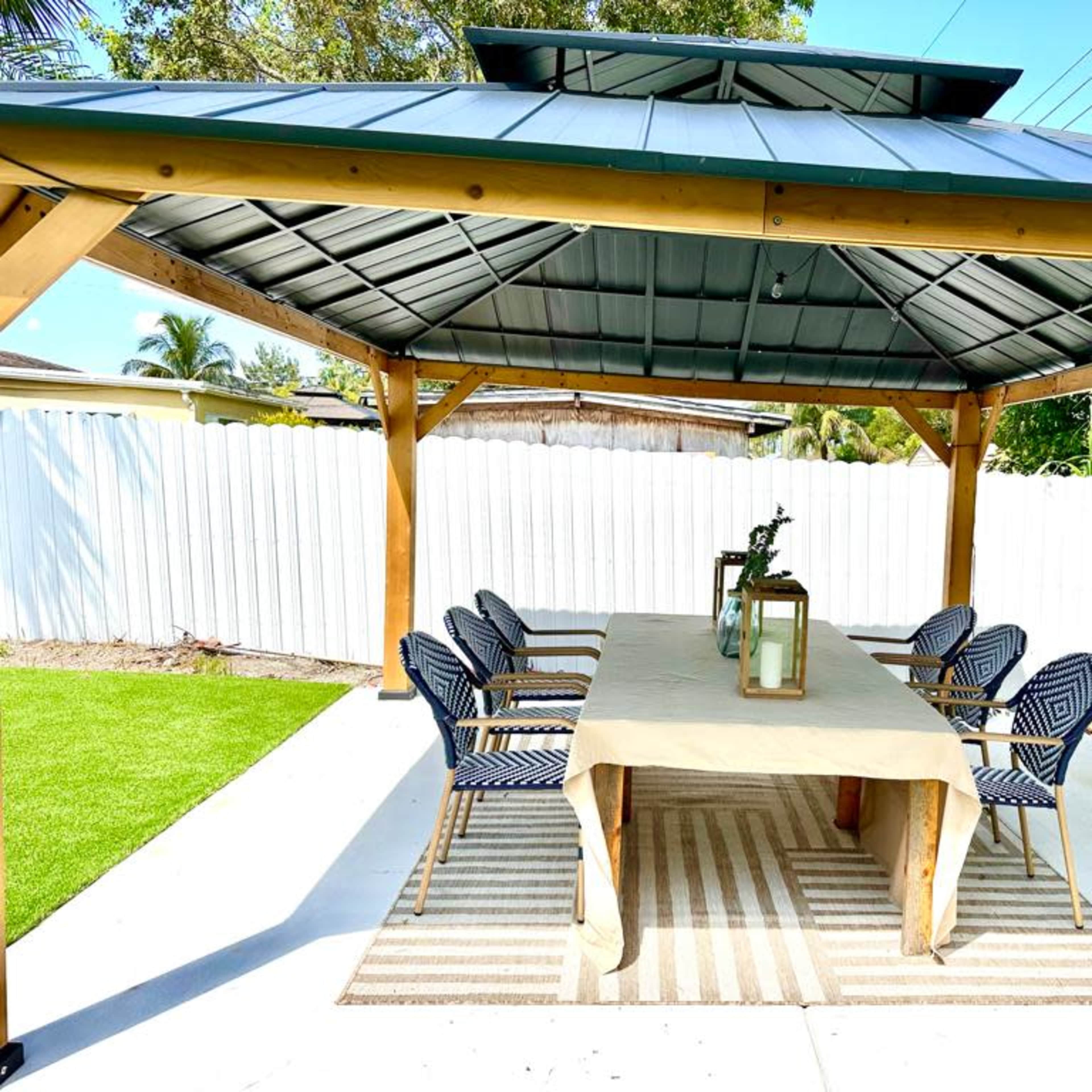 A covered outdoor seating area features a long table with chairs under a wooden gazebo, surrounded by a landscaped yard.