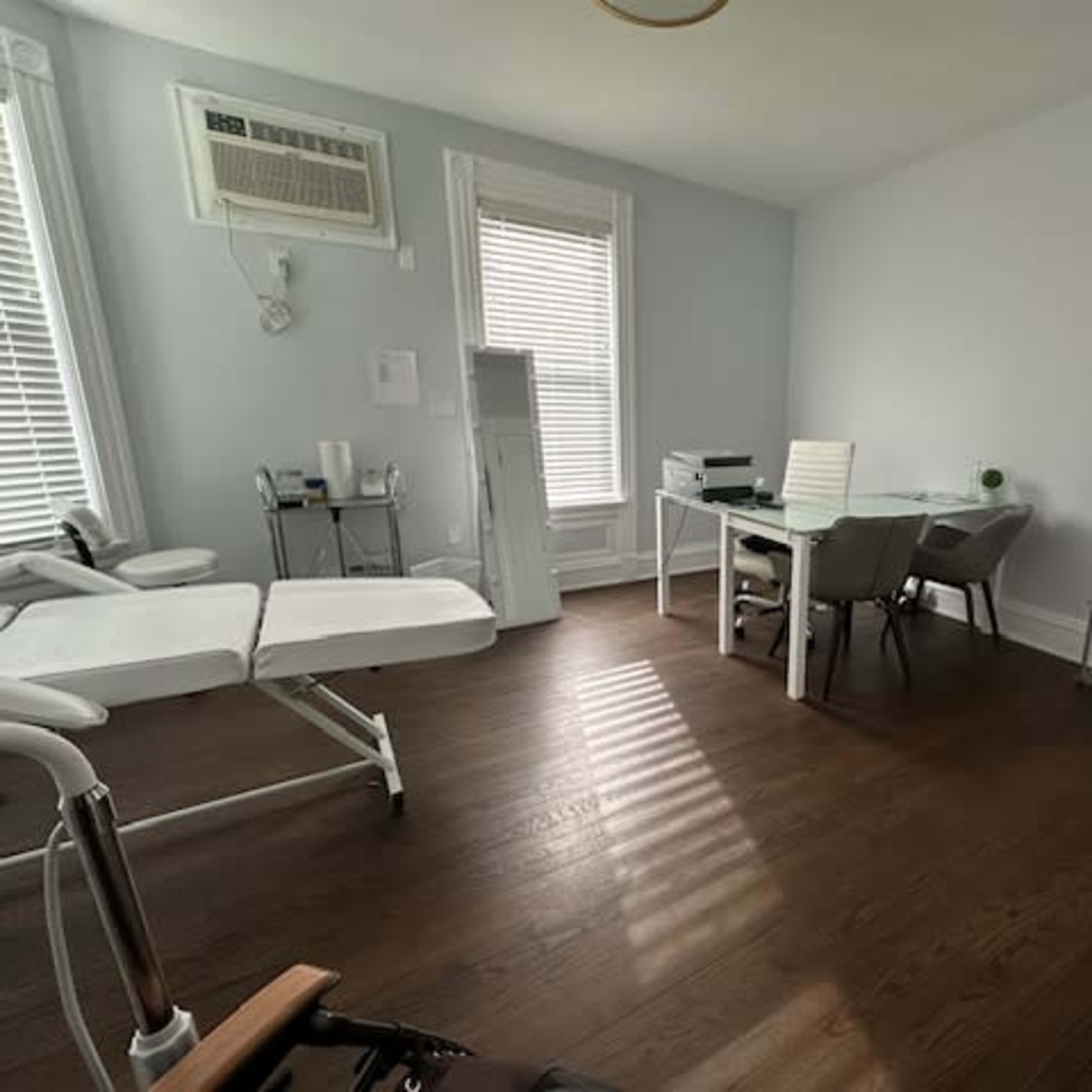 A bright, minimalist office space with a treatment table, a desk with chairs, and a shelving unit.
