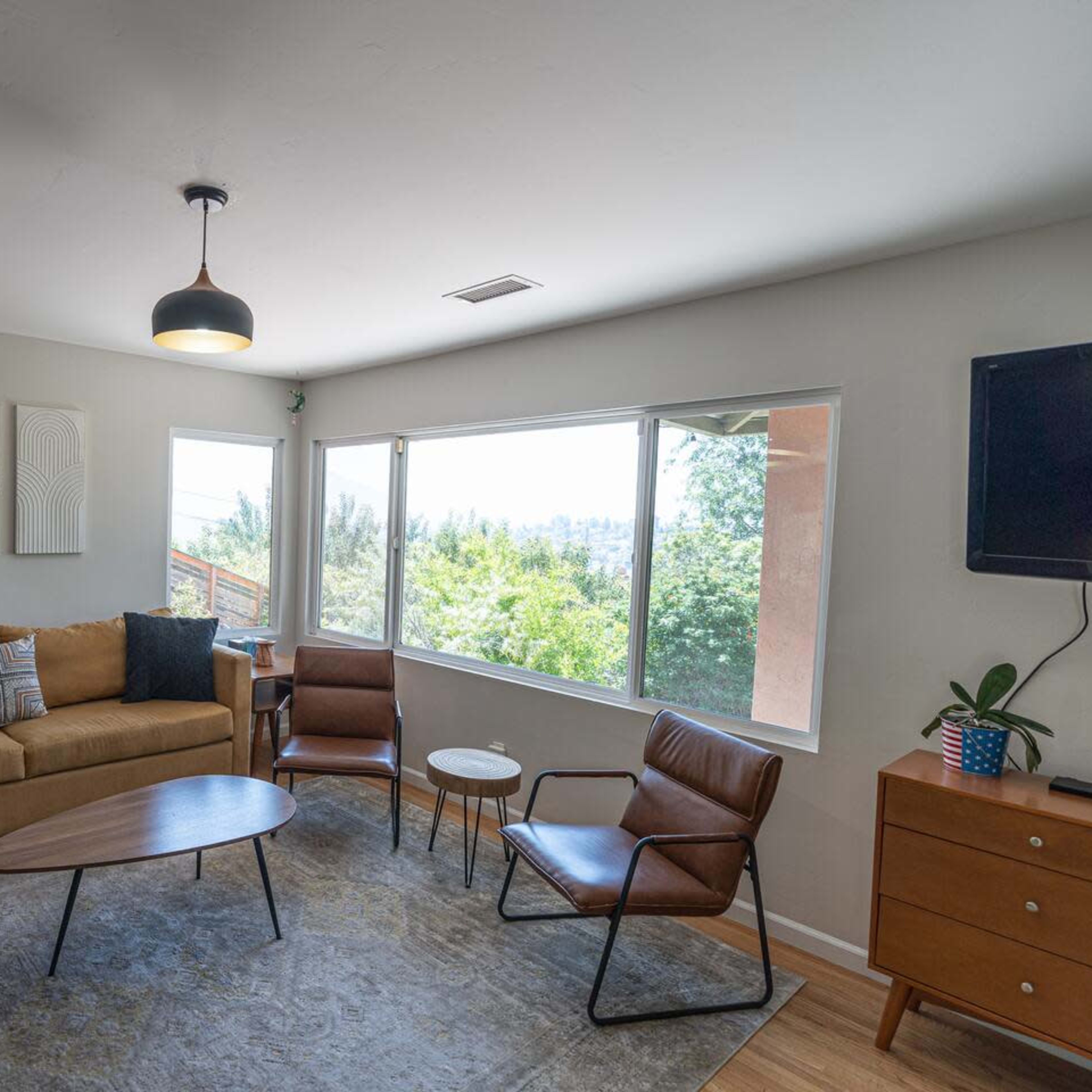 A cozy living room features a brown sofa, a round coffee table, two chairs, and large windows that provide a view of greenery outside.