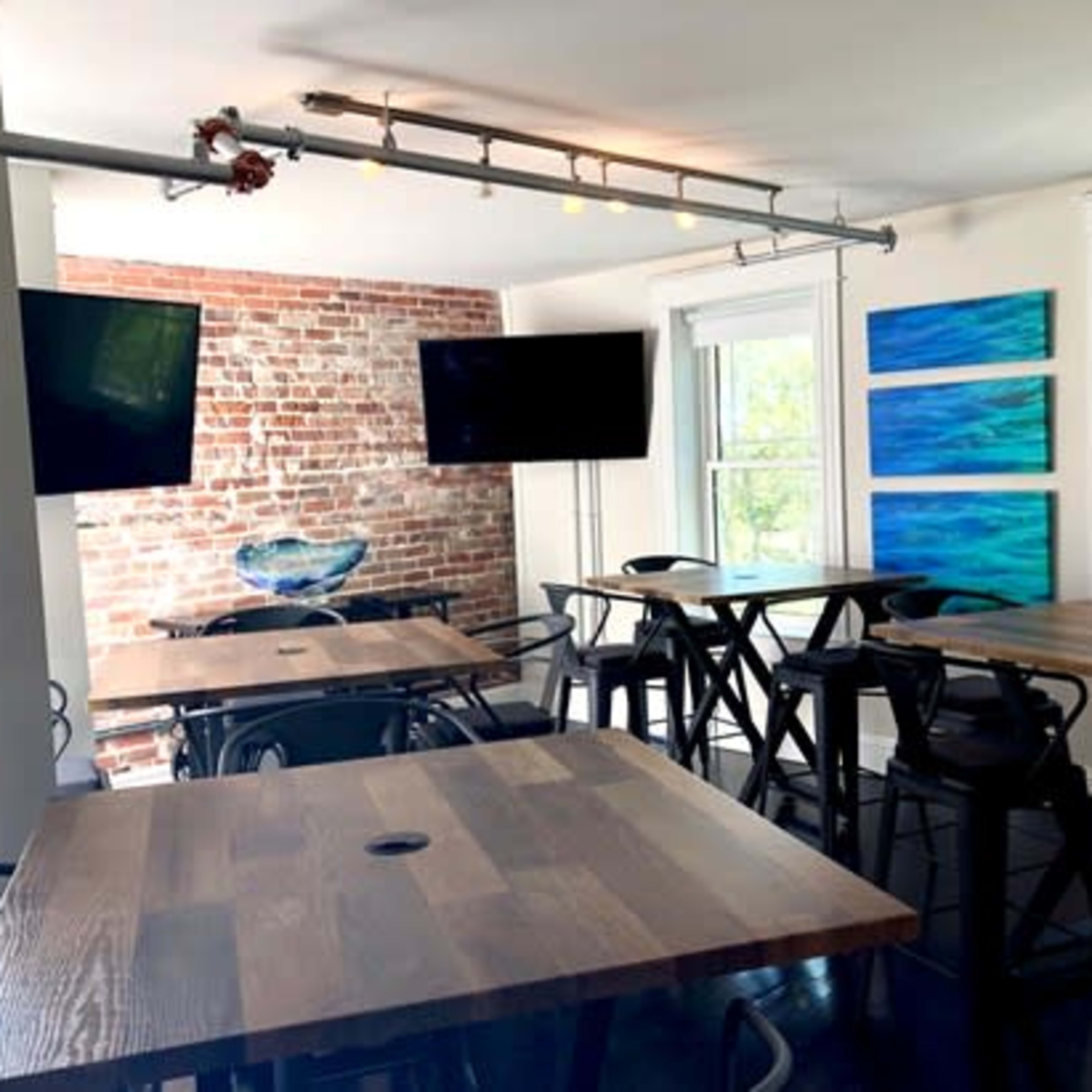 A modern room featuring several wooden tables, two wall-mounted televisions, and abstract blue artworks on the walls, with a brick accent wall.