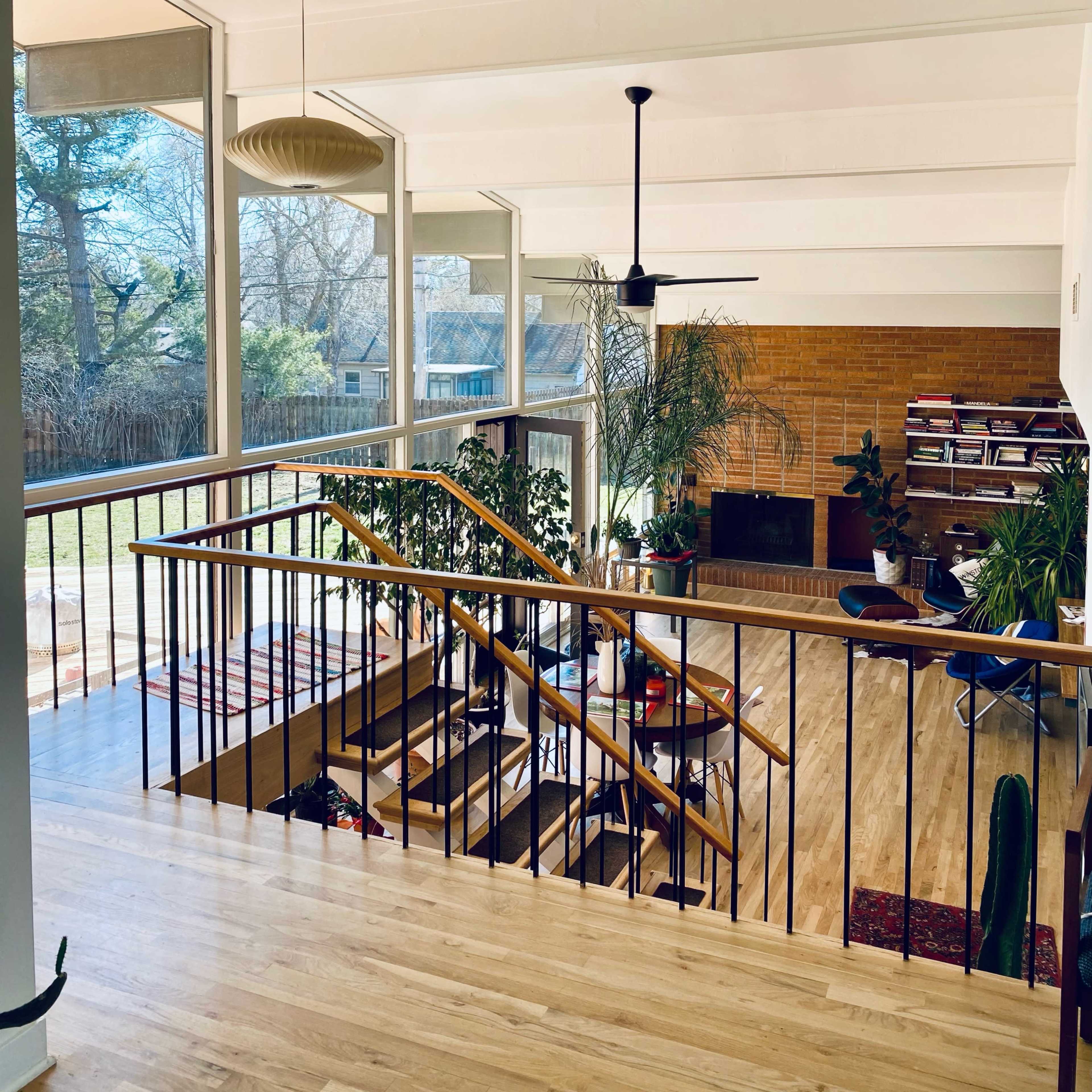 A spacious, modern living area with a wooden staircase, large windows, and plants, leading to a patio and backyard.
