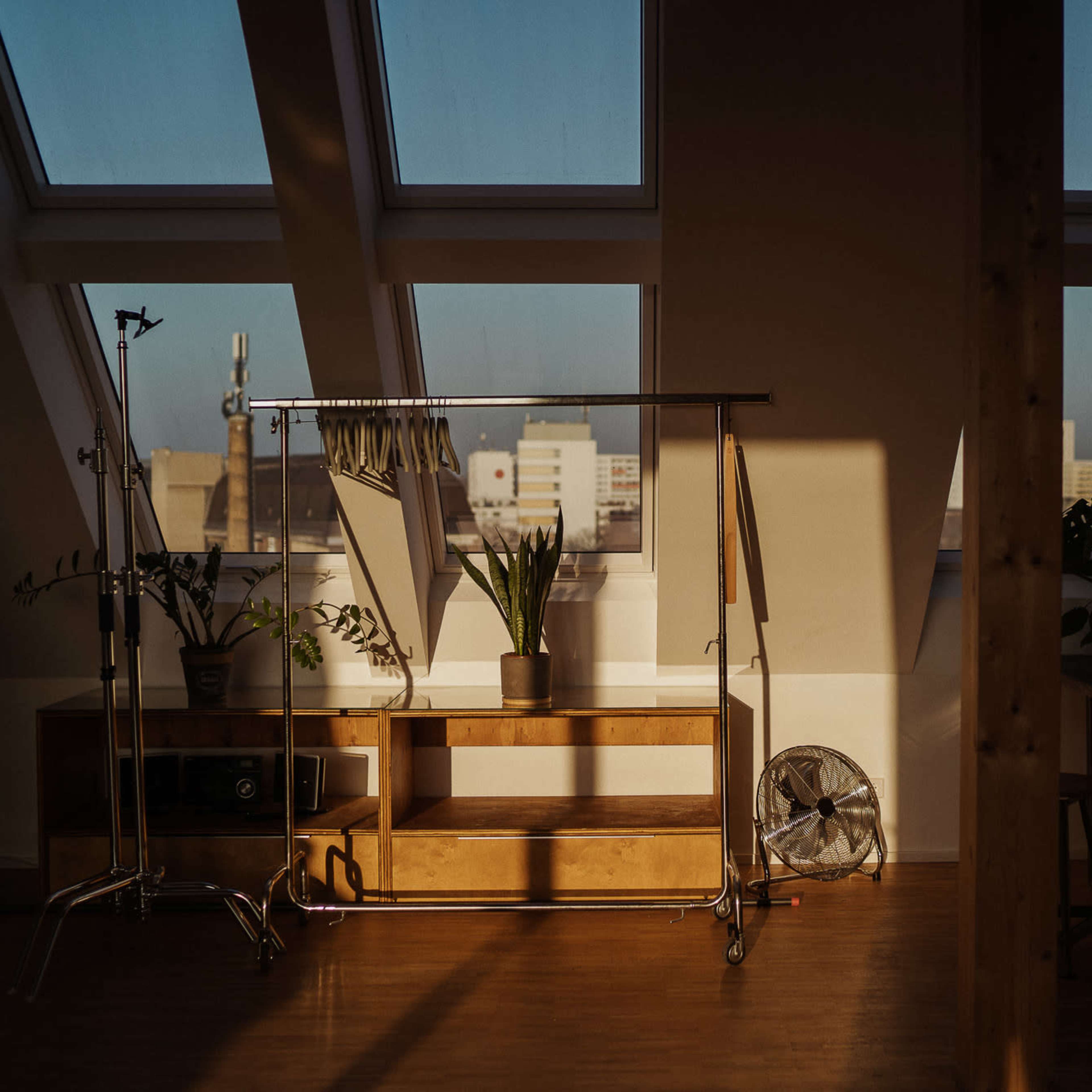 An airy attic interior with large windows, a wooden clothing rack, a shelf with plants, and a fan, casting shadows on the floor.