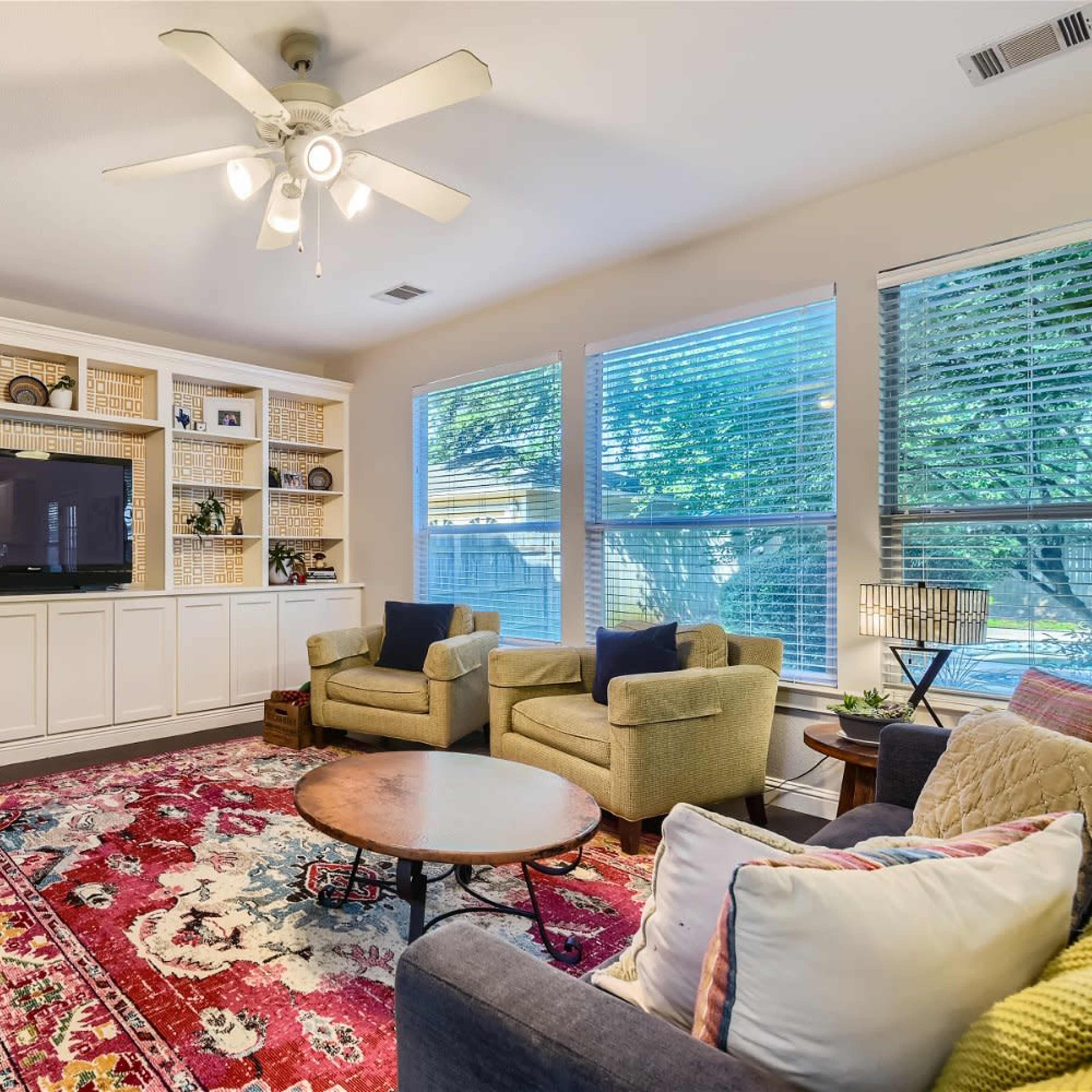 A living room with a large flat-screen TV mounted above a built-in shelf, two armchairs, and a coffee table on a colorful area rug.
