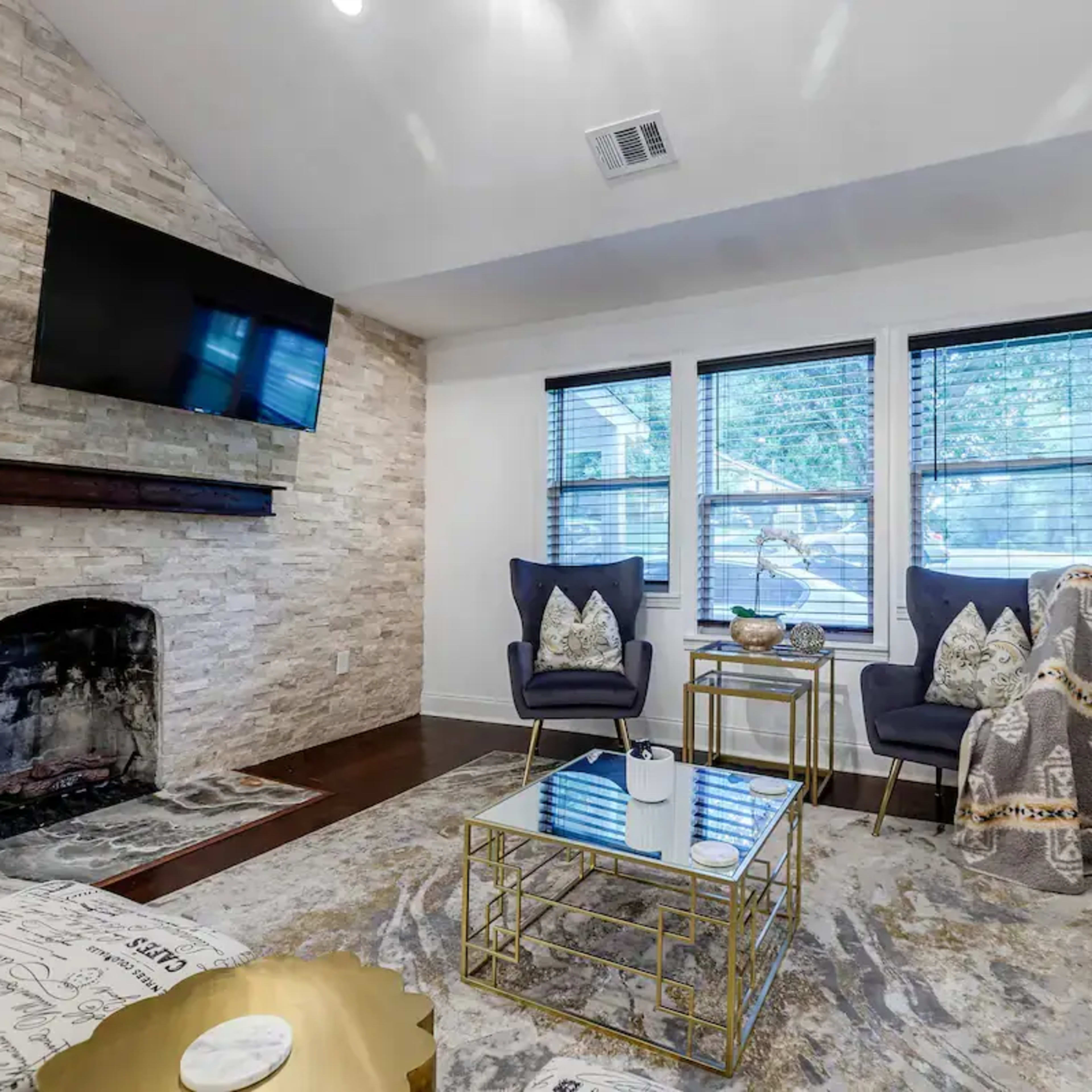 A living room features a stone accent wall with a mounted TV, two armchairs, a coffee table, and large windows allowing natural light to enter.