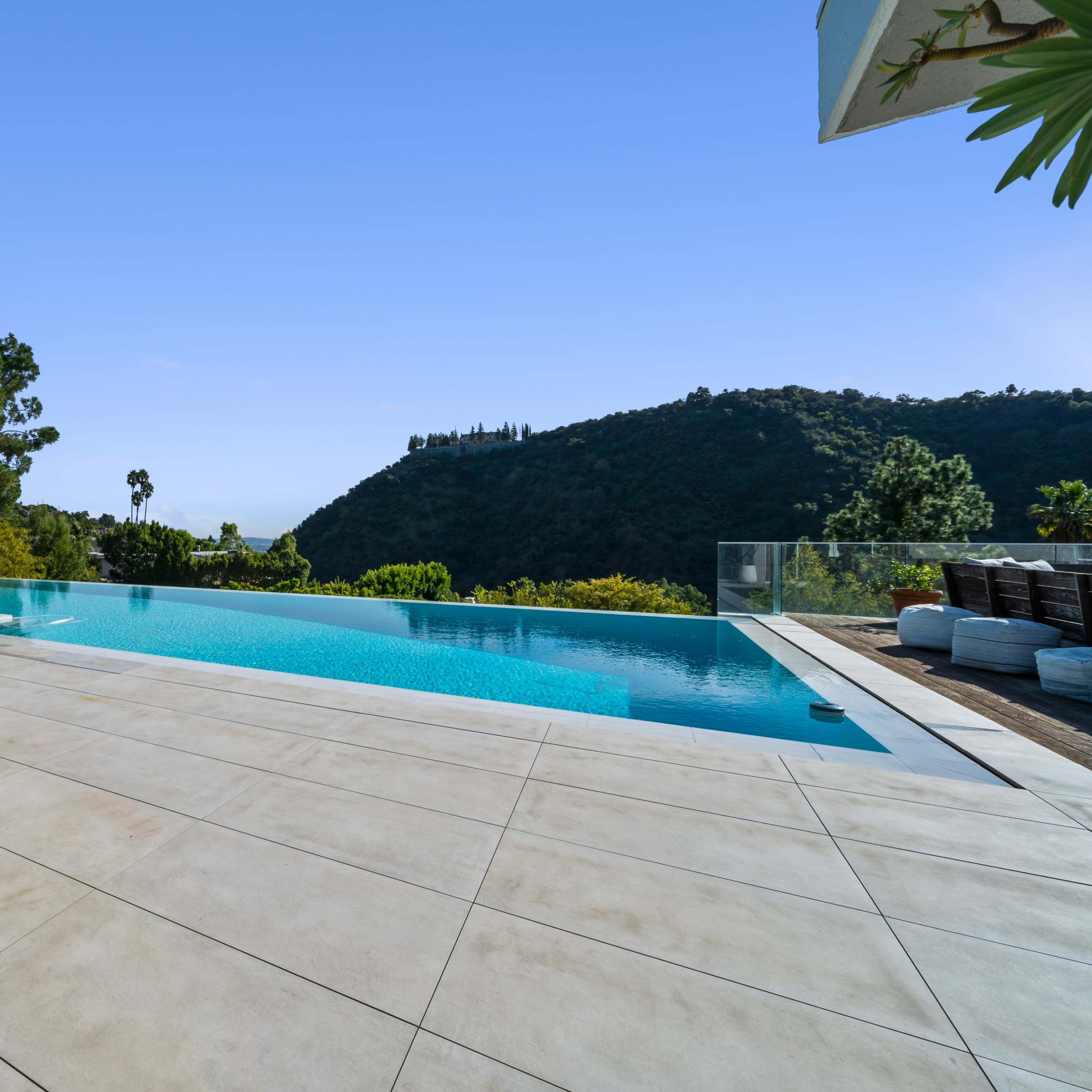 A modern outdoor pool area surrounded by lounge chairs and umbrellas, with a hillside view in the background.