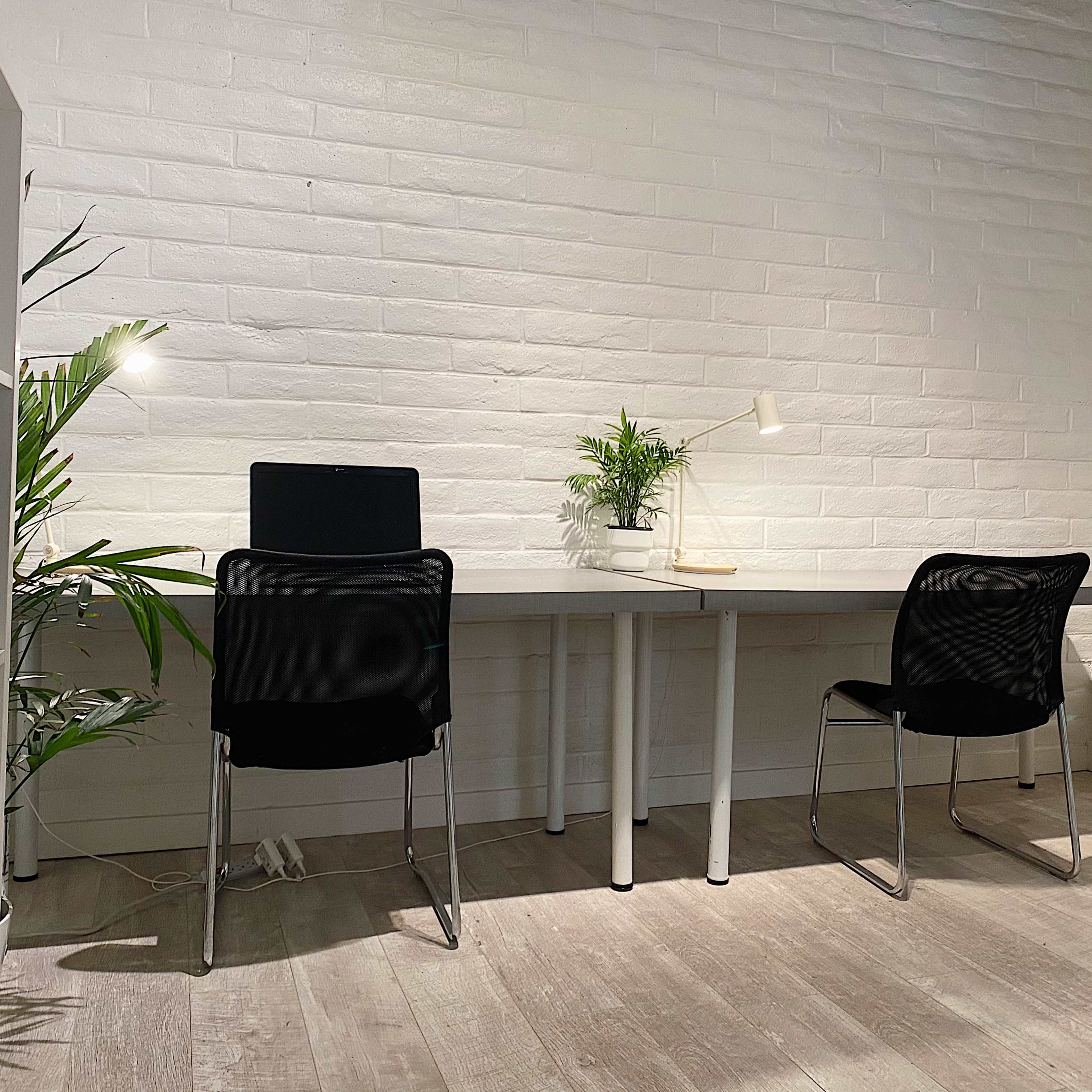 A minimalistic workspace features two black chairs at a light gray table, with a small plant and a lamp on the table against a white brick wall.