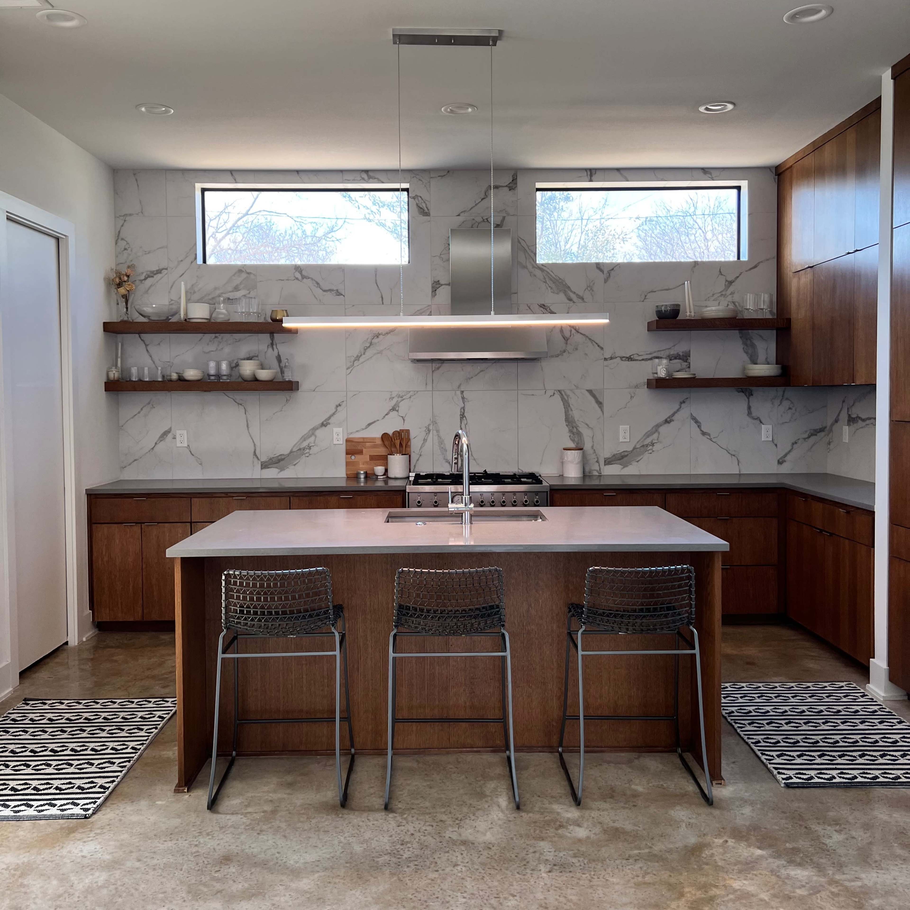 A modern kitchen featuring a central island with three metal chairs, dark wood cabinetry, and marble-patterned backsplash.