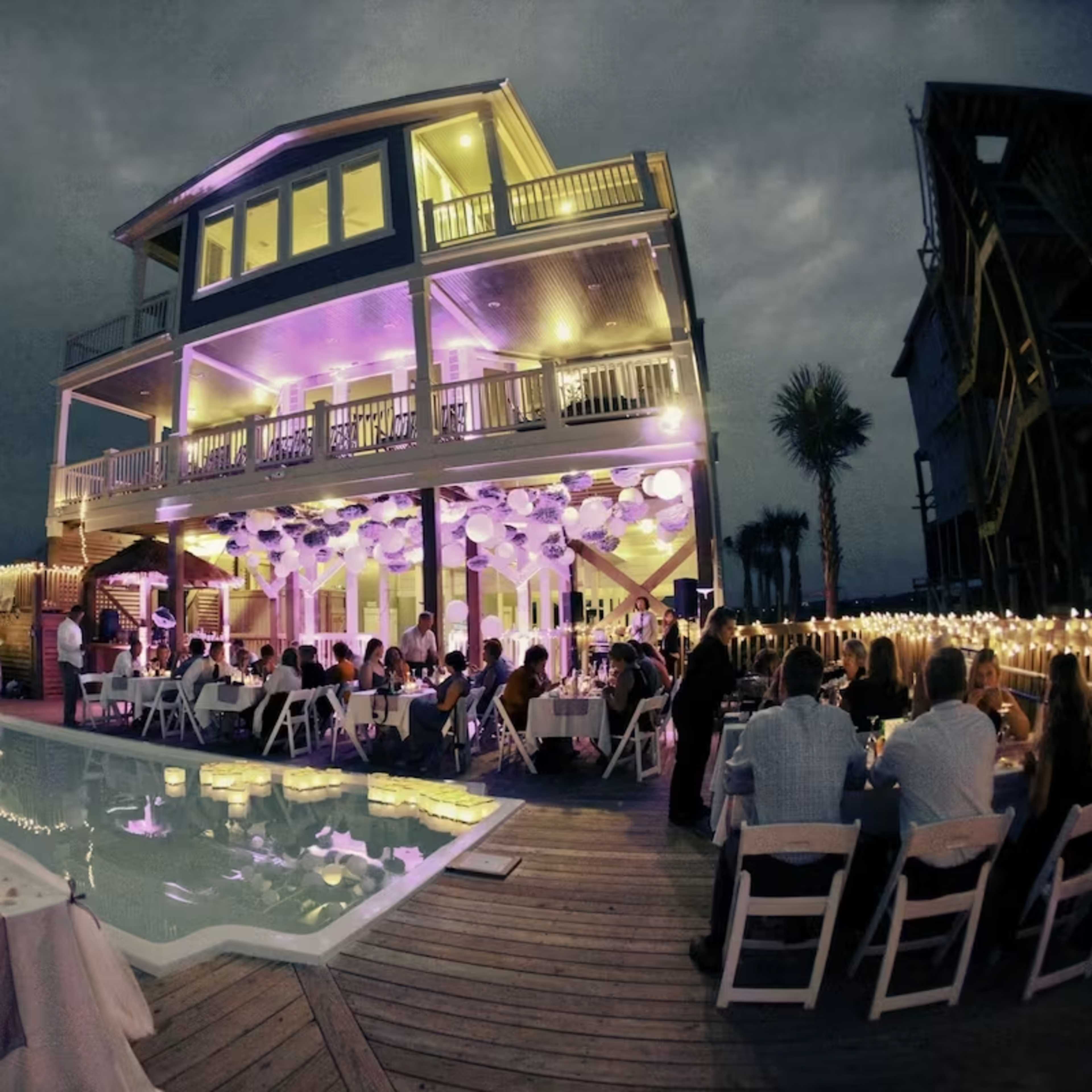 A large gathering is taking place around a pool at a beautifully lit house, decorated with string lights and purple accents, as guests sit at tables.