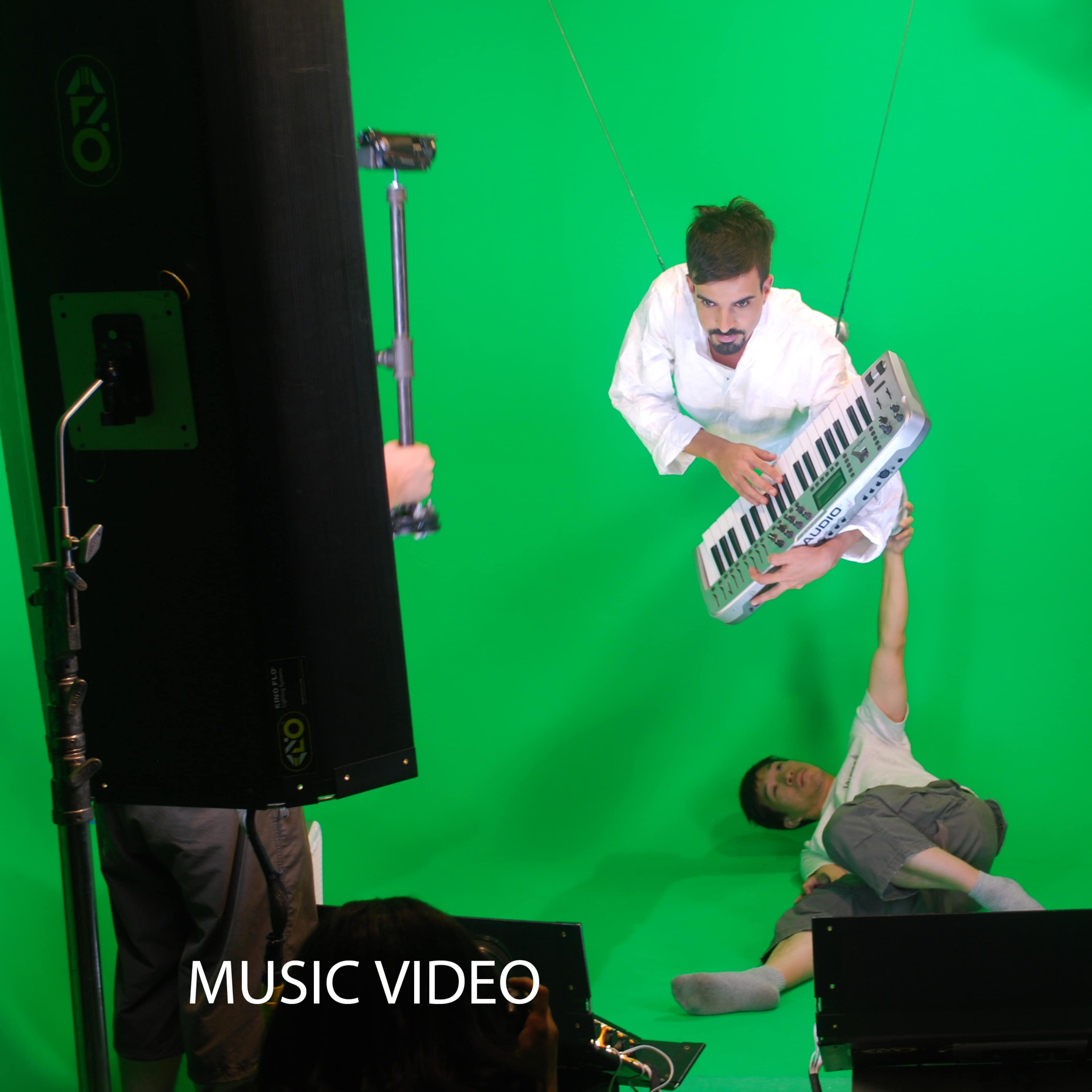 A man in a white outfit plays a keyboard while suspended above another man lying on the ground in a green screen studio setting.