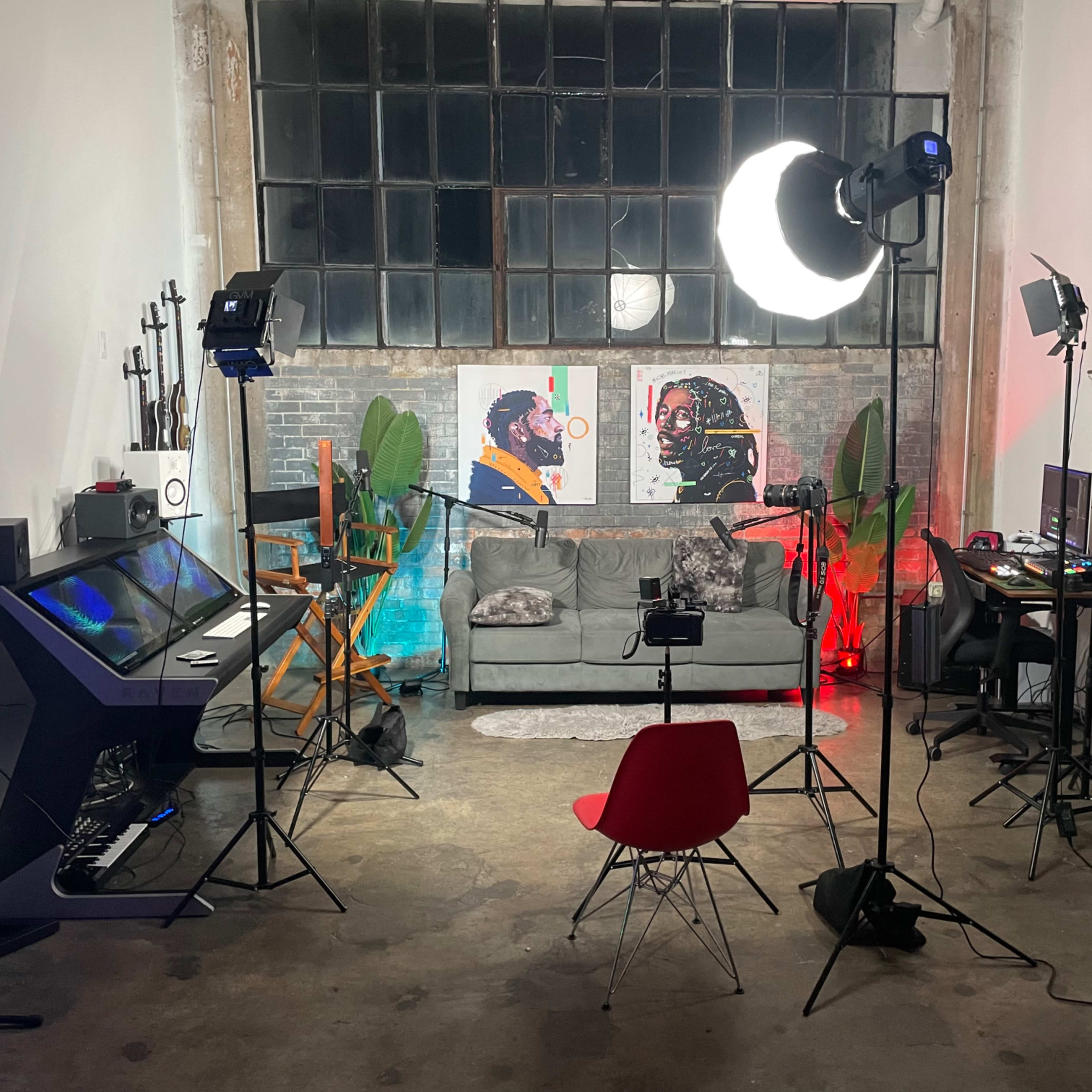 A well-lit studio space with two art pieces on the wall, a gray couch in the center, and equipment set up for video production, including cameras and lights.