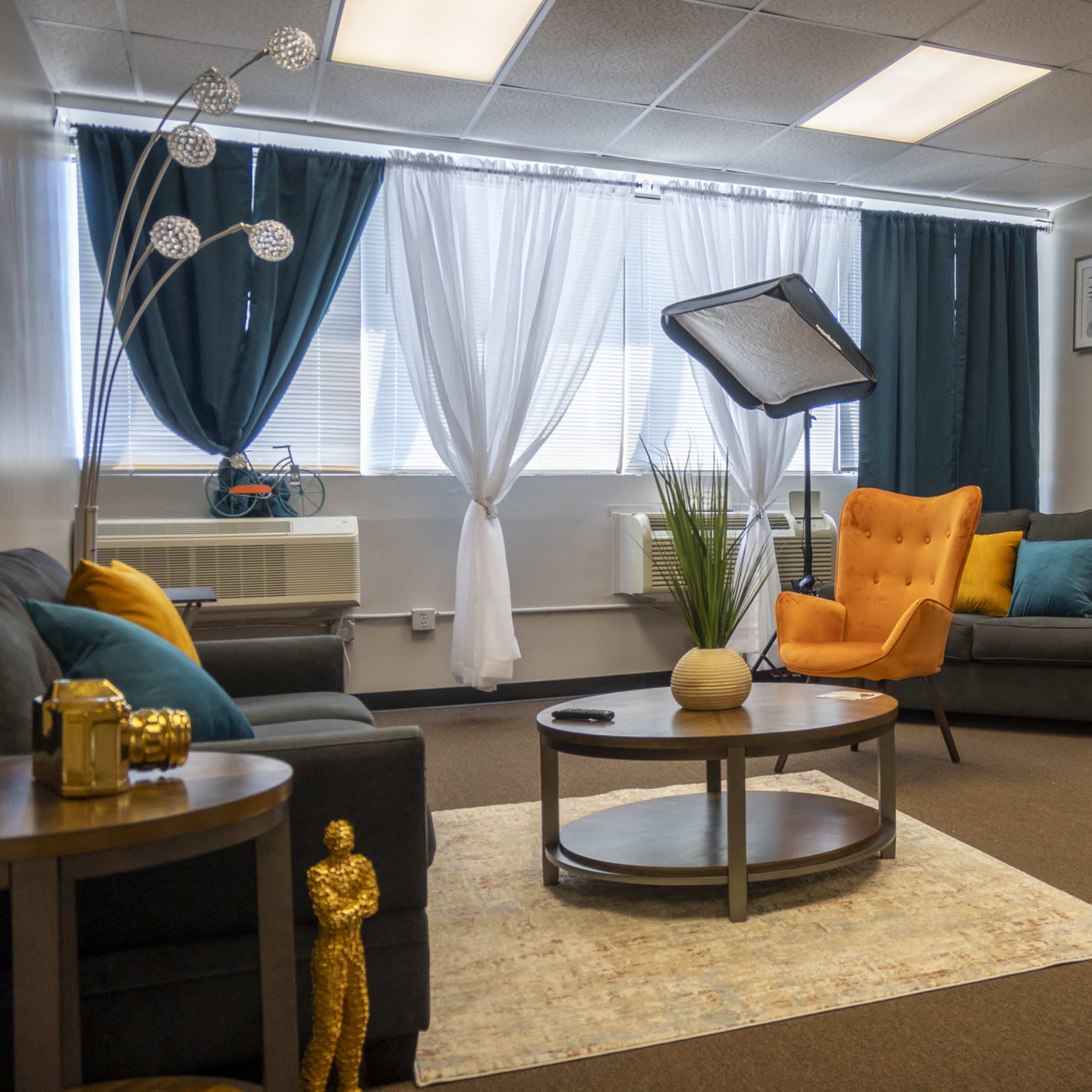 A modern office lounge area with two orange chairs, a round coffee table, and a large window dressed in light curtains.