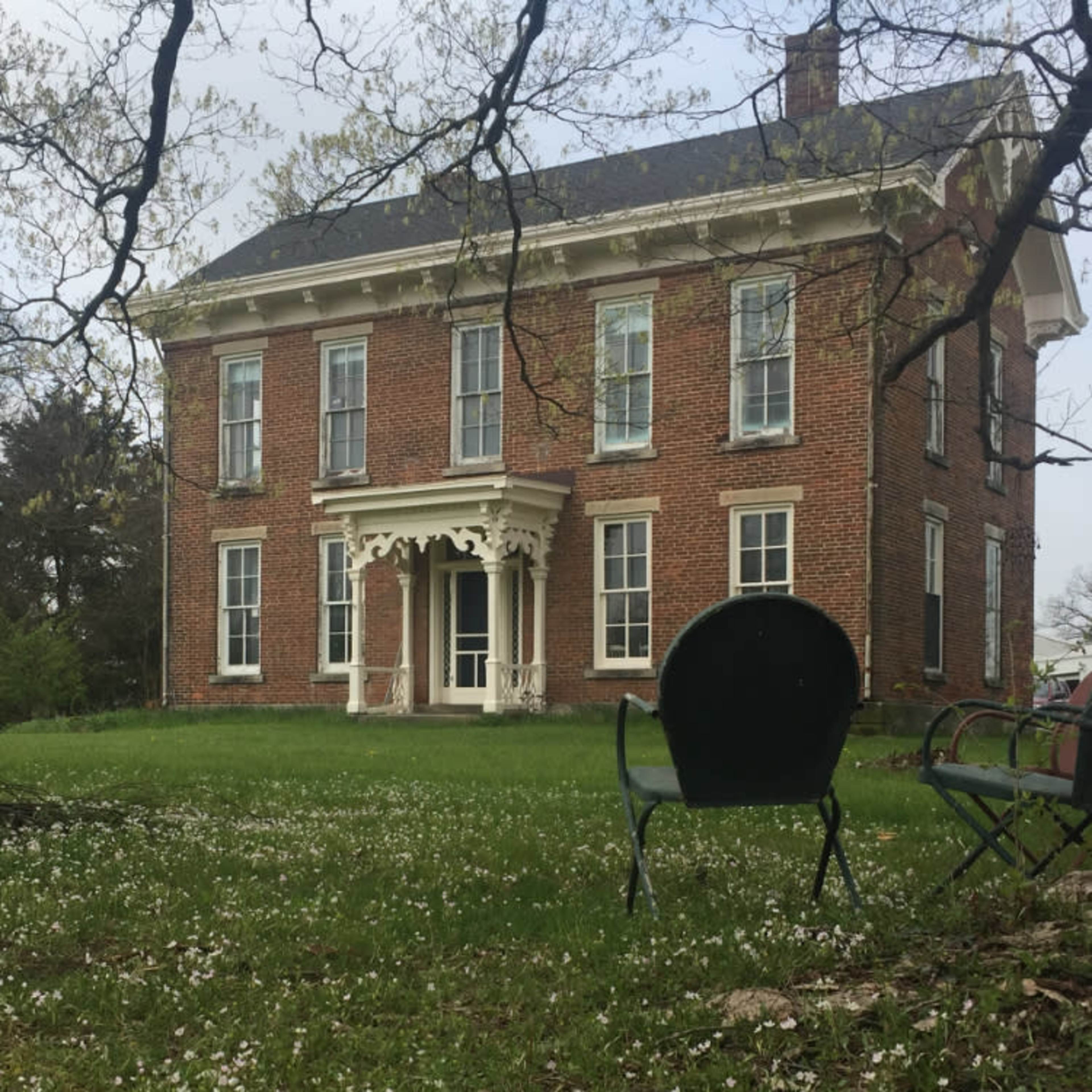 A large brick house with white trim stands in a grassy yard scattered with white flowers, and two green chairs are positioned in the foreground.