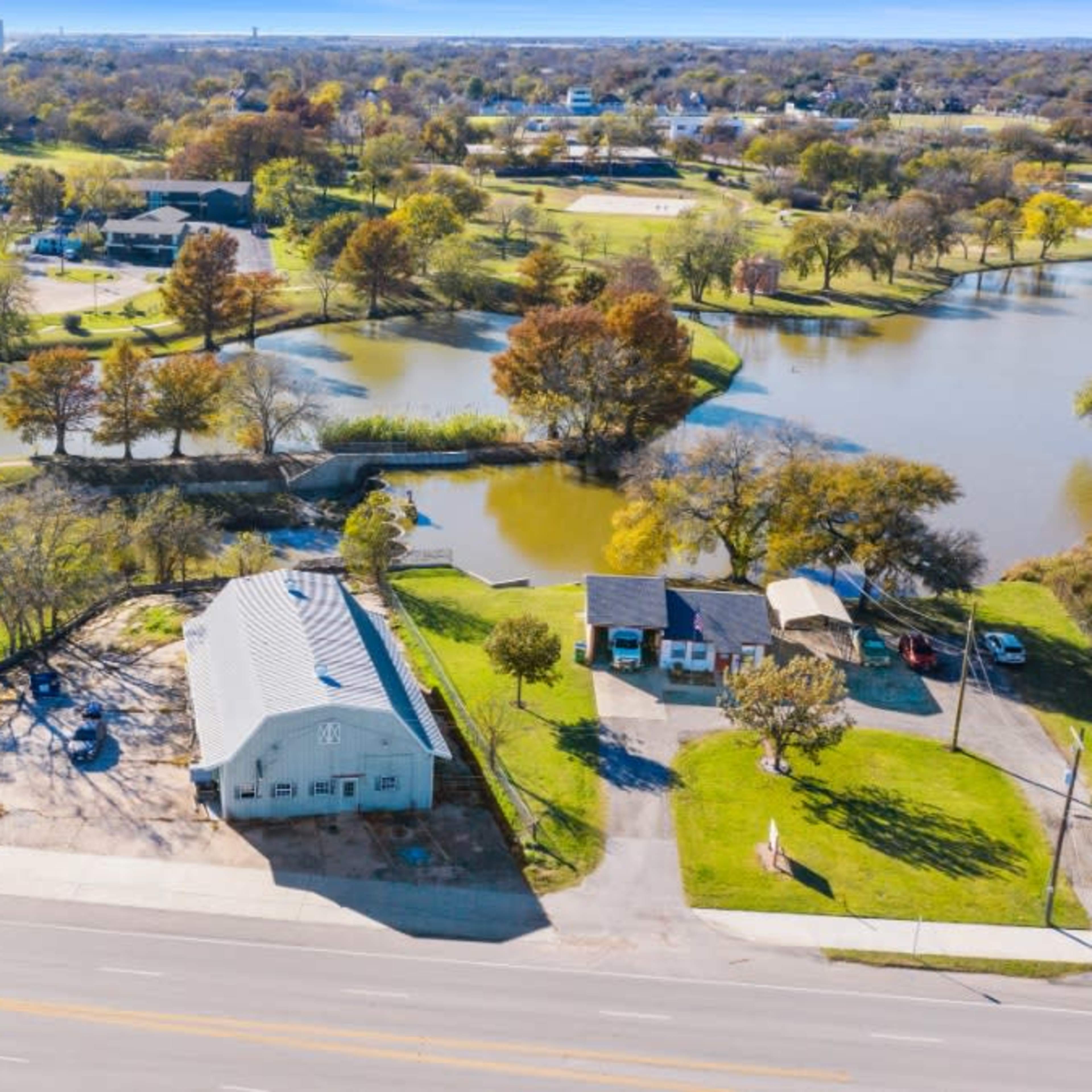 An aerial view of a landscape featuring a house near a water body, with several trees and a park-like area in the background.