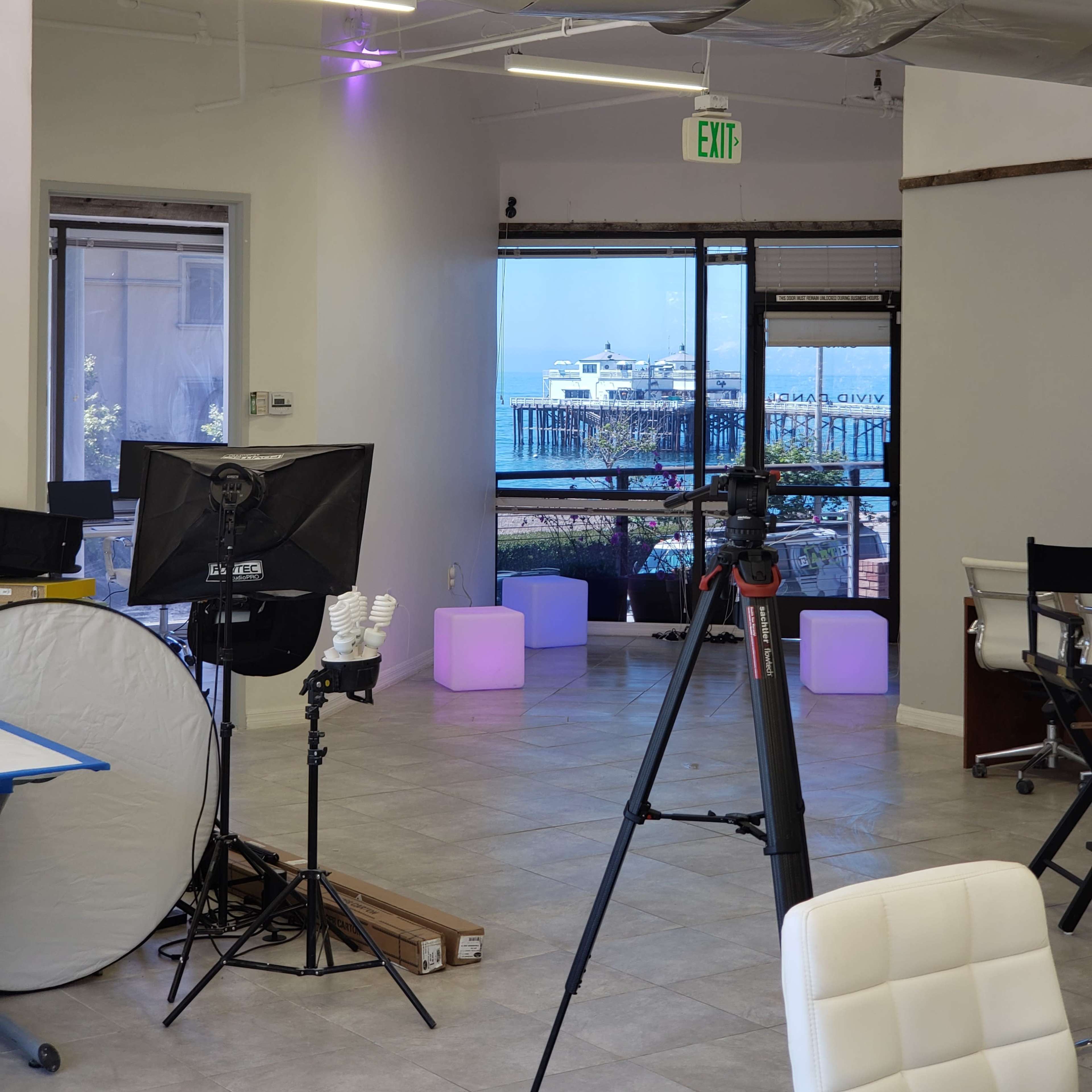 An office space with a view of a pier through large windows, featuring modern lighting and photography equipment set up inside.