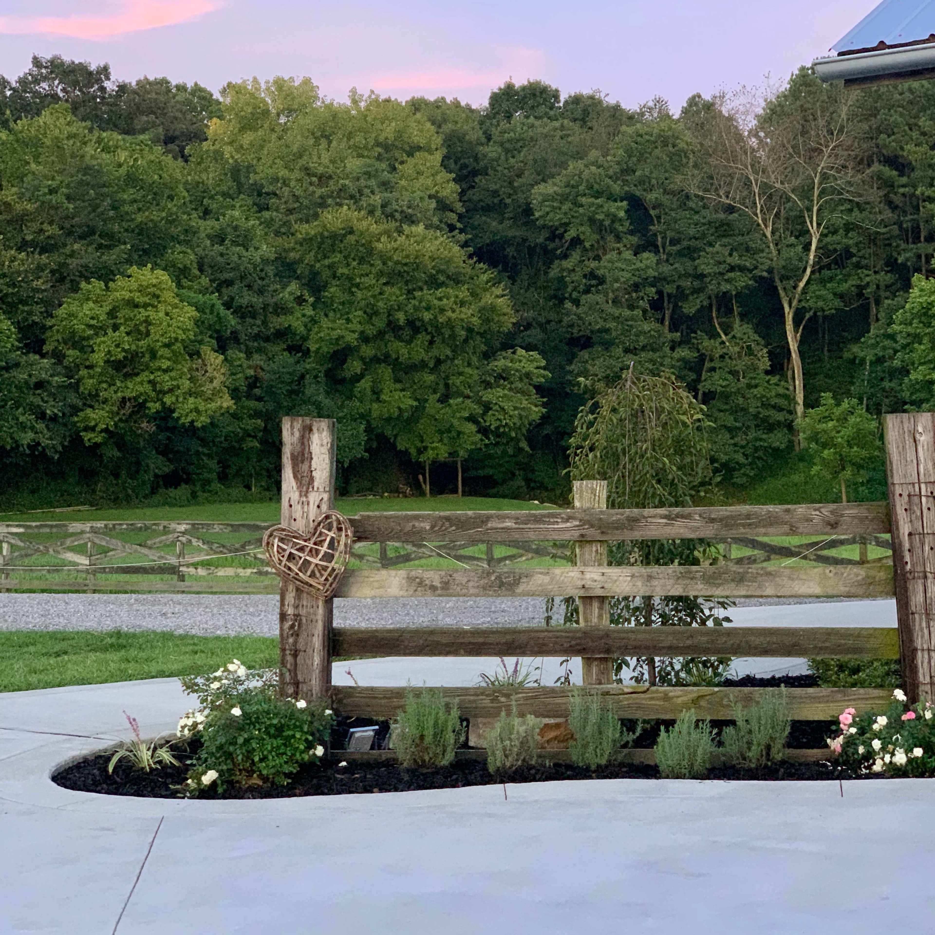 A wooden fence surrounds a garden with flowers in the foreground, set against a backdrop of greenery and a twilight sky.