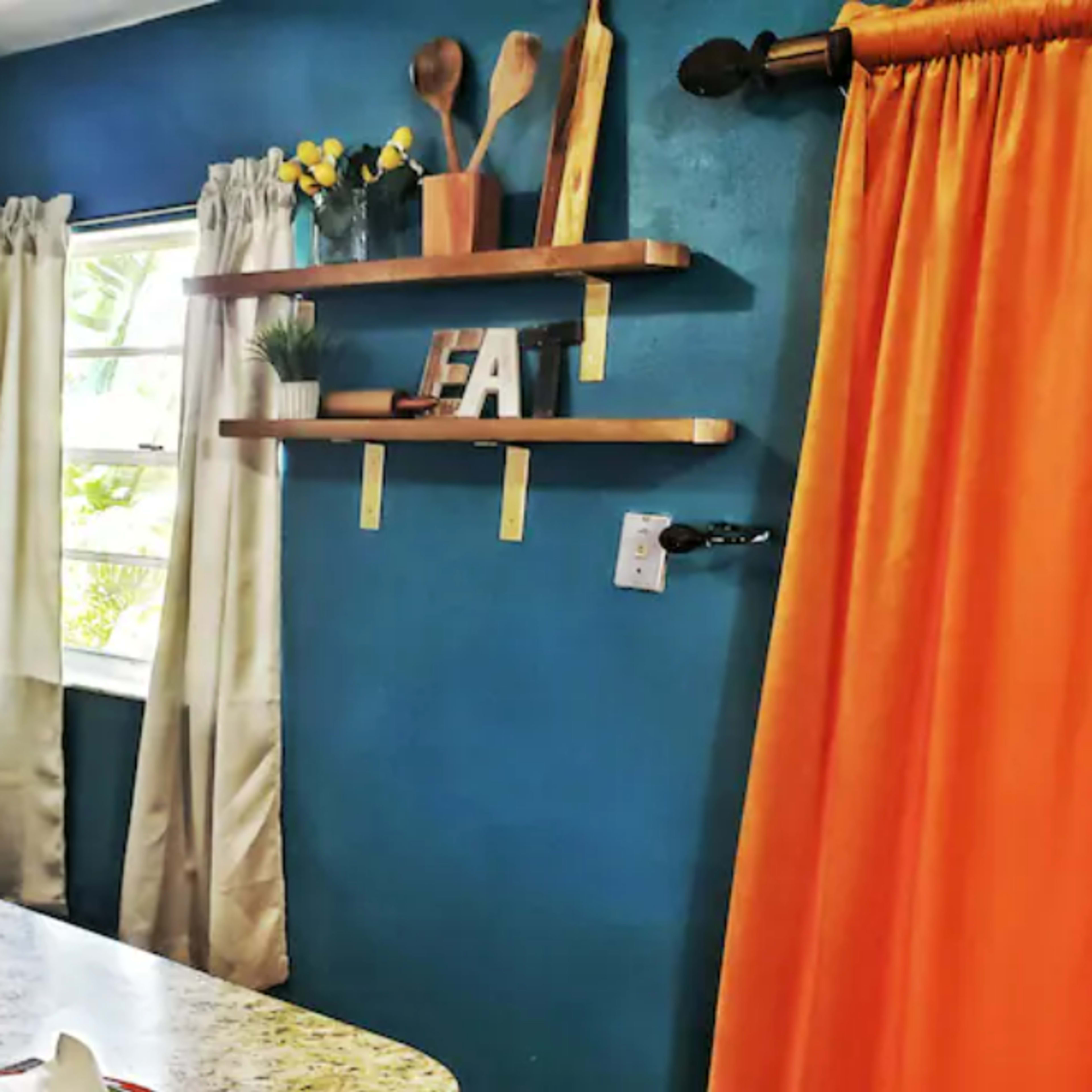 A bright kitchen corner features a refrigerator, a door, and a shelf adorned with kitchen utensils, while an orange curtain frames a window.