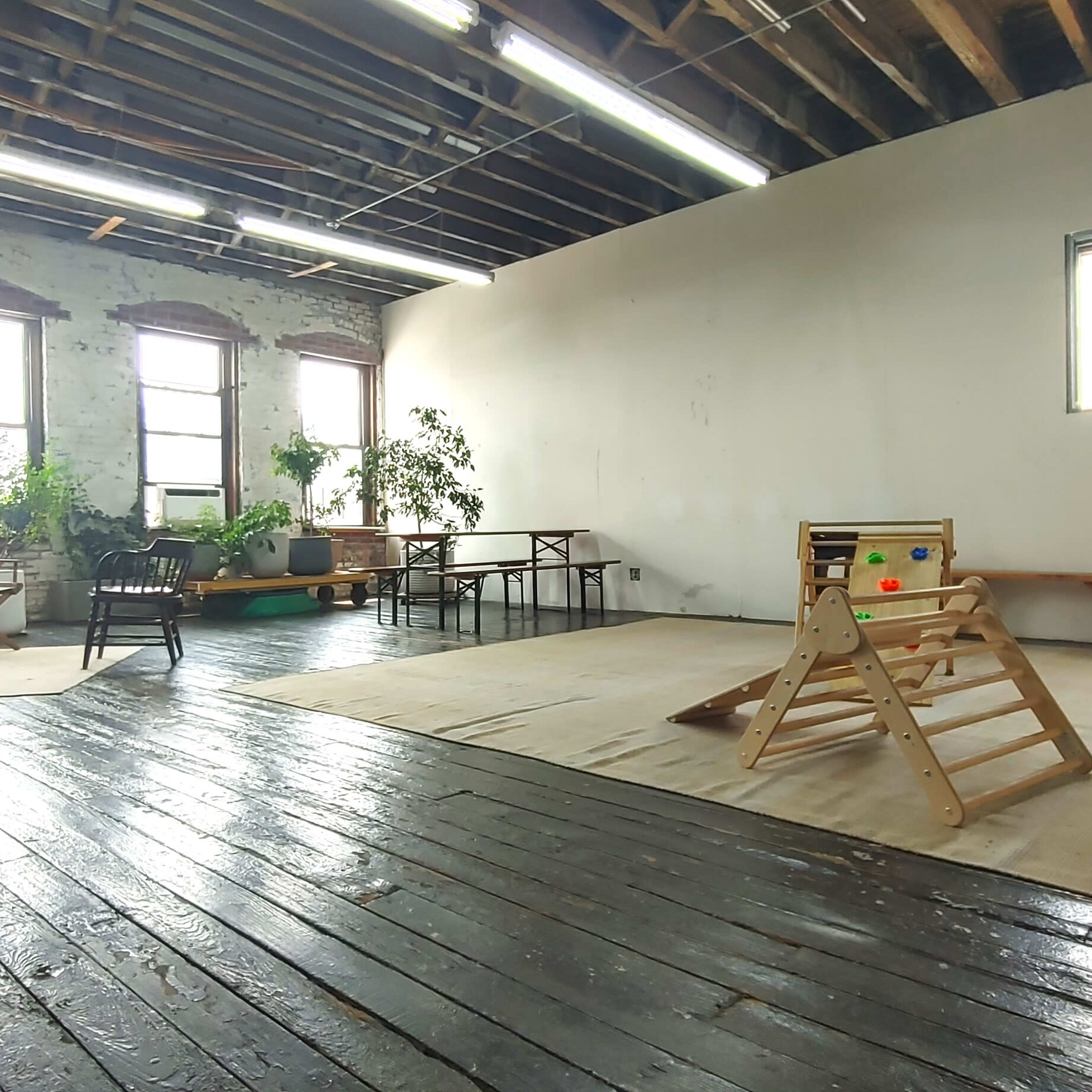 A spacious room with wooden floors, large windows, and minimal furnishings, including a climbing structure and scattered toys.