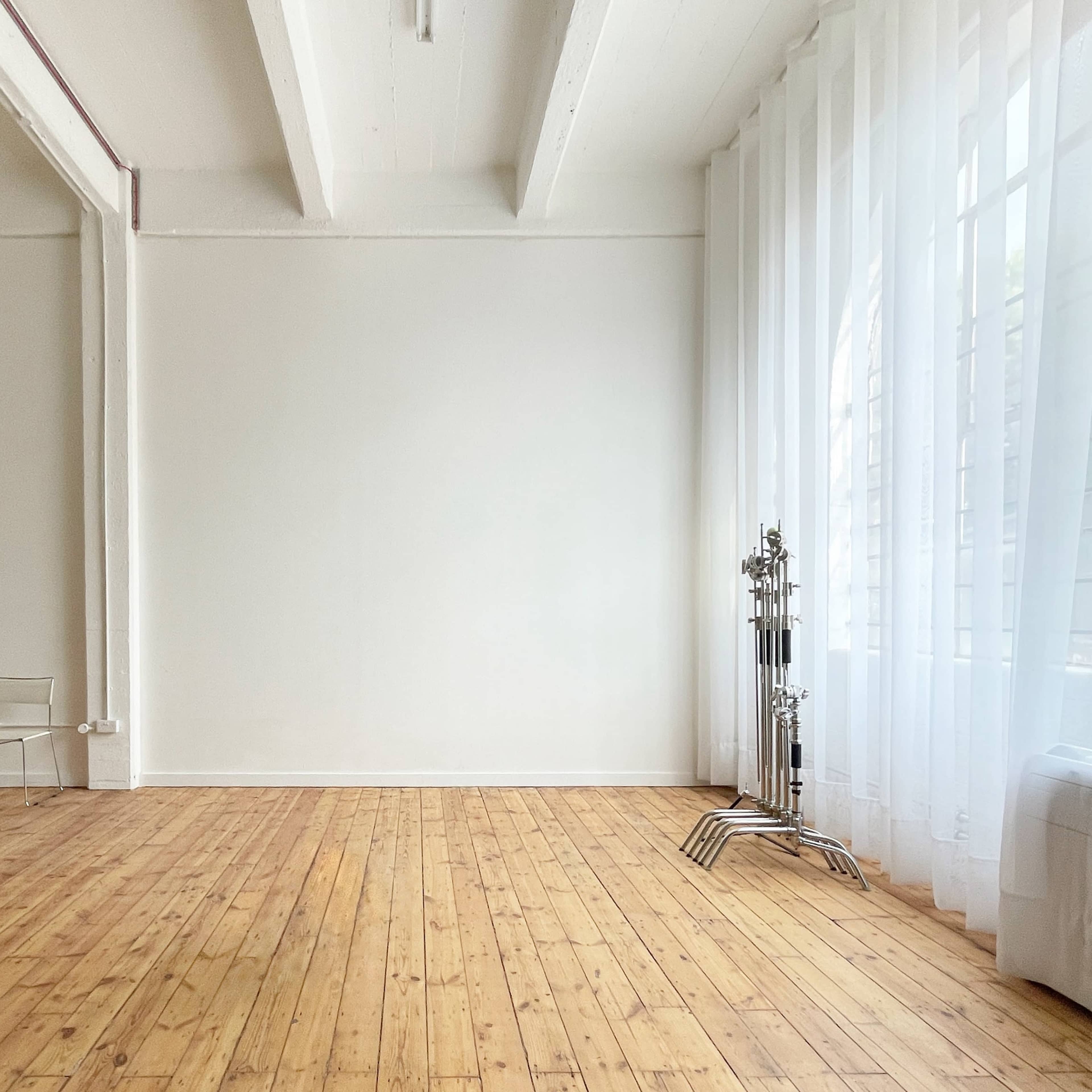 A spacious, minimalist interior with wooden flooring, large windows covered by sheer curtains, and a few pieces of furniture including chairs and a decorative stand.