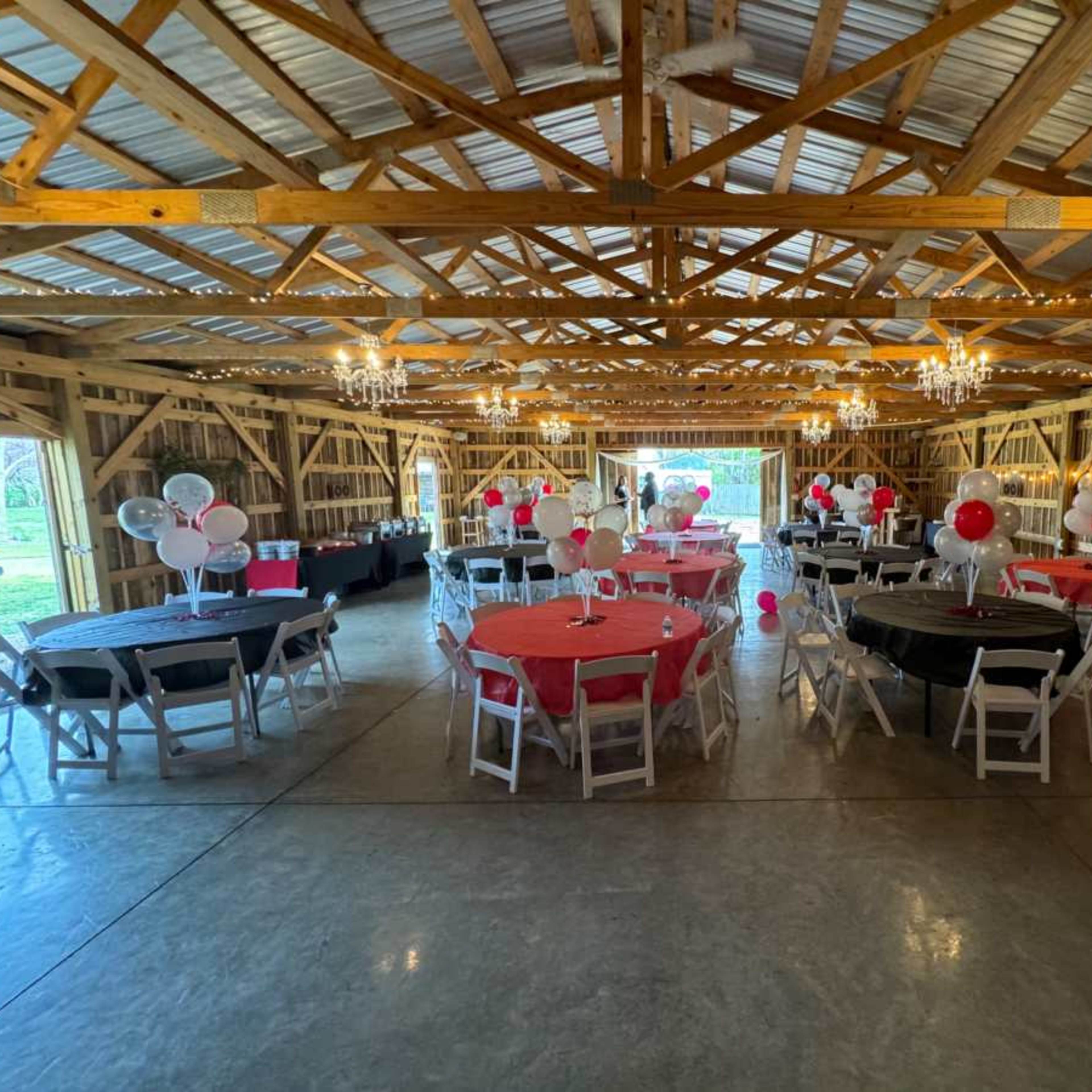 The interior of a barn is set up for a party, featuring round tables covered in red, black, and white linens, with decorations including balloons and chandeliers.