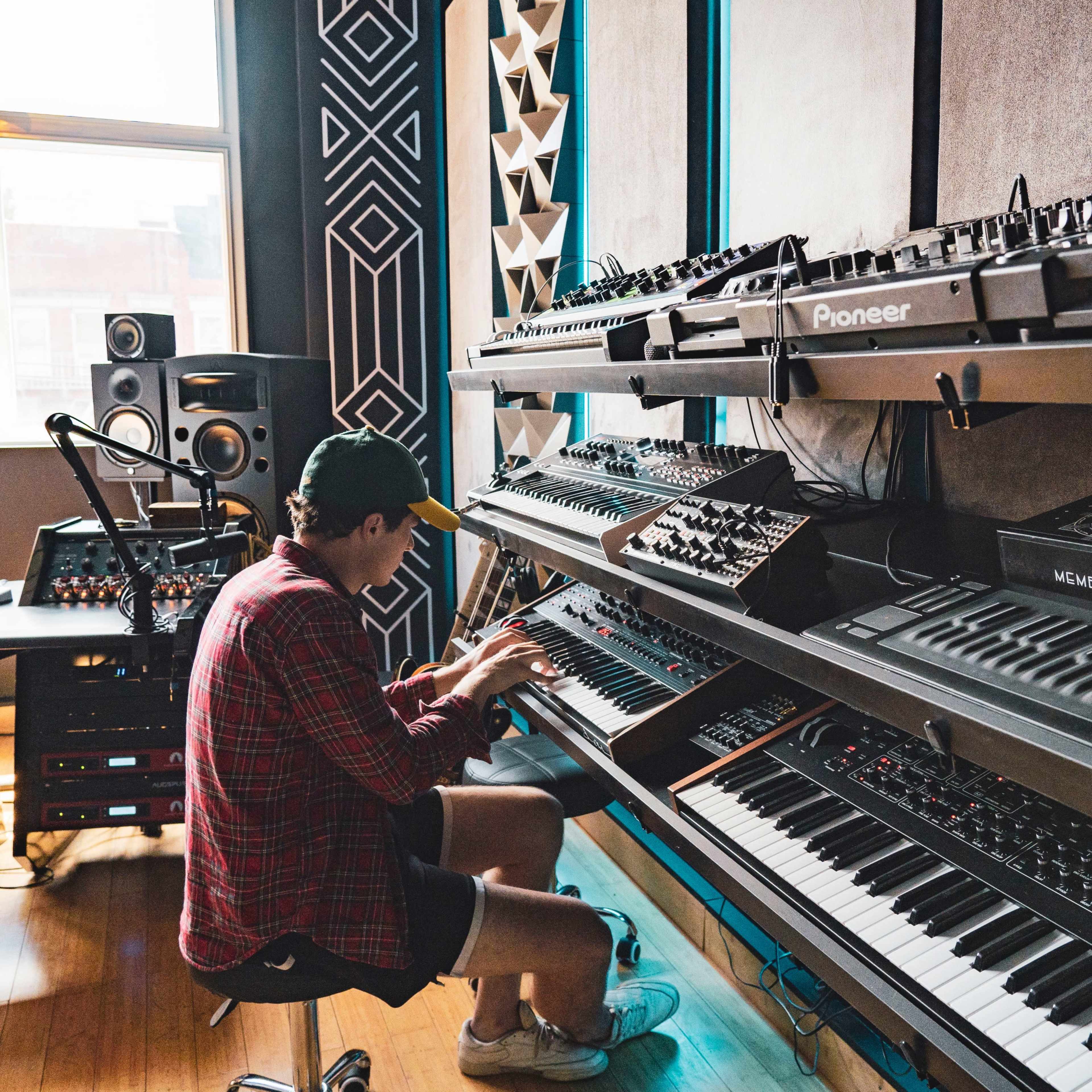 A person wearing a cap is seated on the floor, interacting with a variety of synthesizers and keyboards arranged on metal shelves in a music studio.