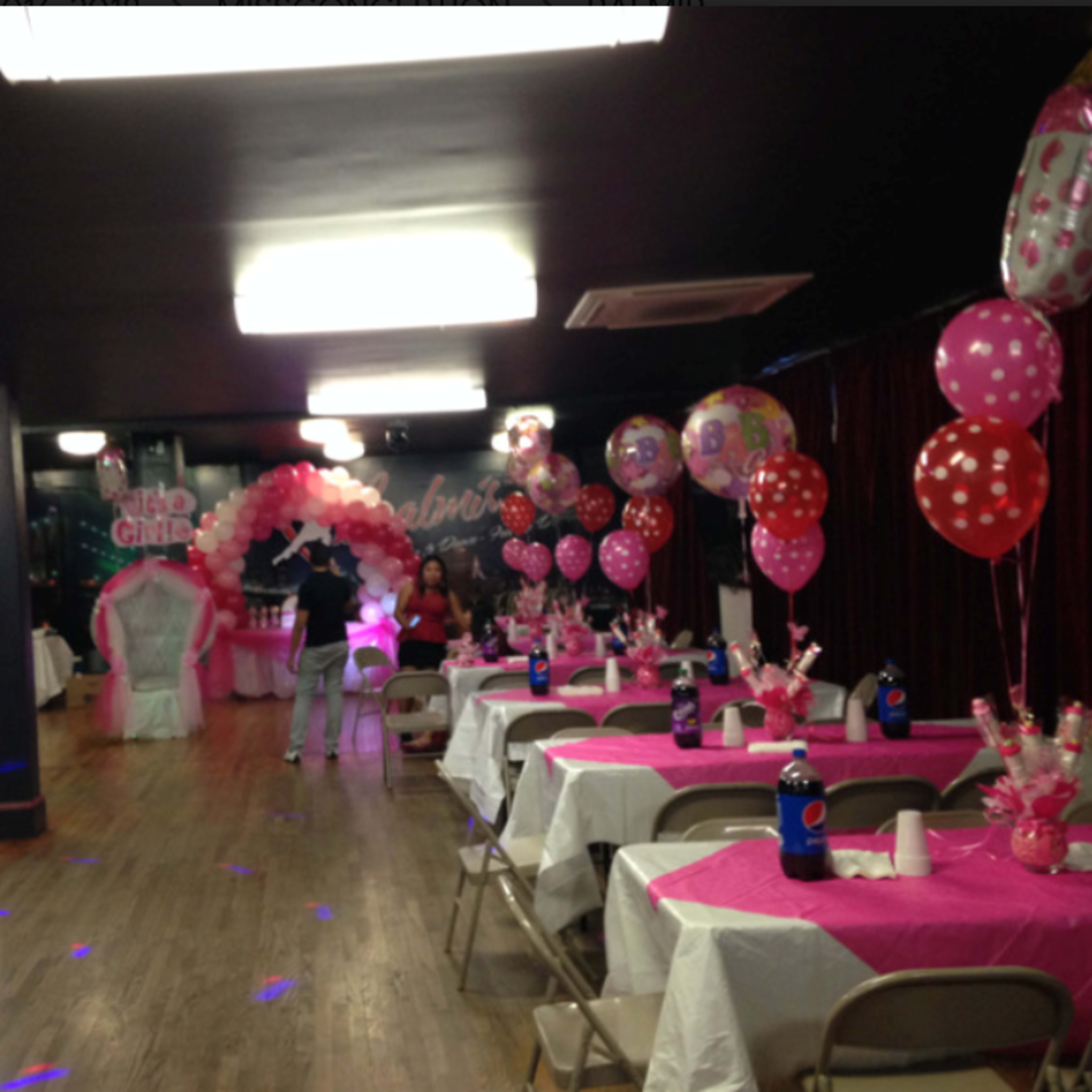 The room is decorated with pink tablecloths, balloons, and a large arch for a celebration.