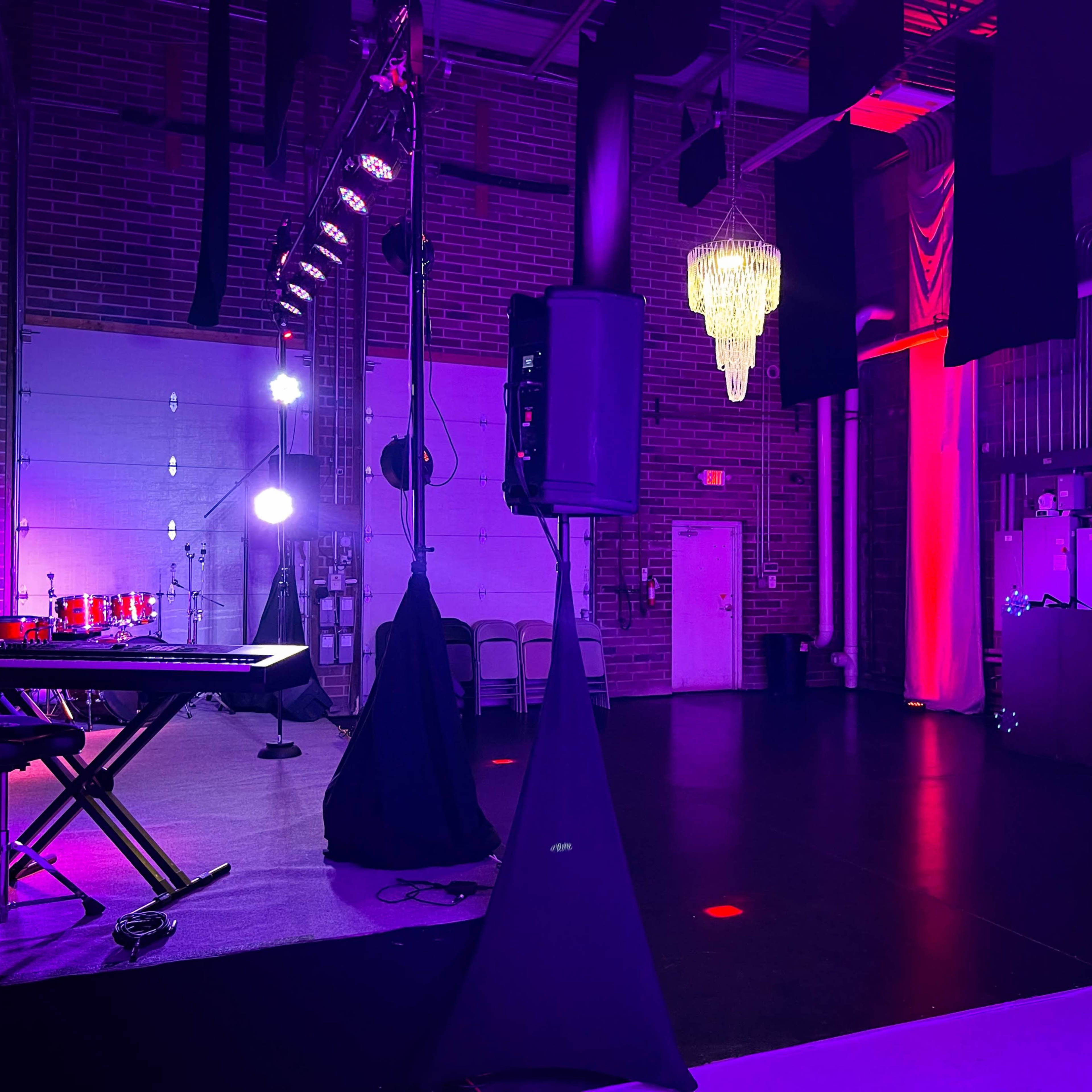 A dimly lit event space with a stage featuring a keyboard and a chandelier, surrounded by purple and red lighting.