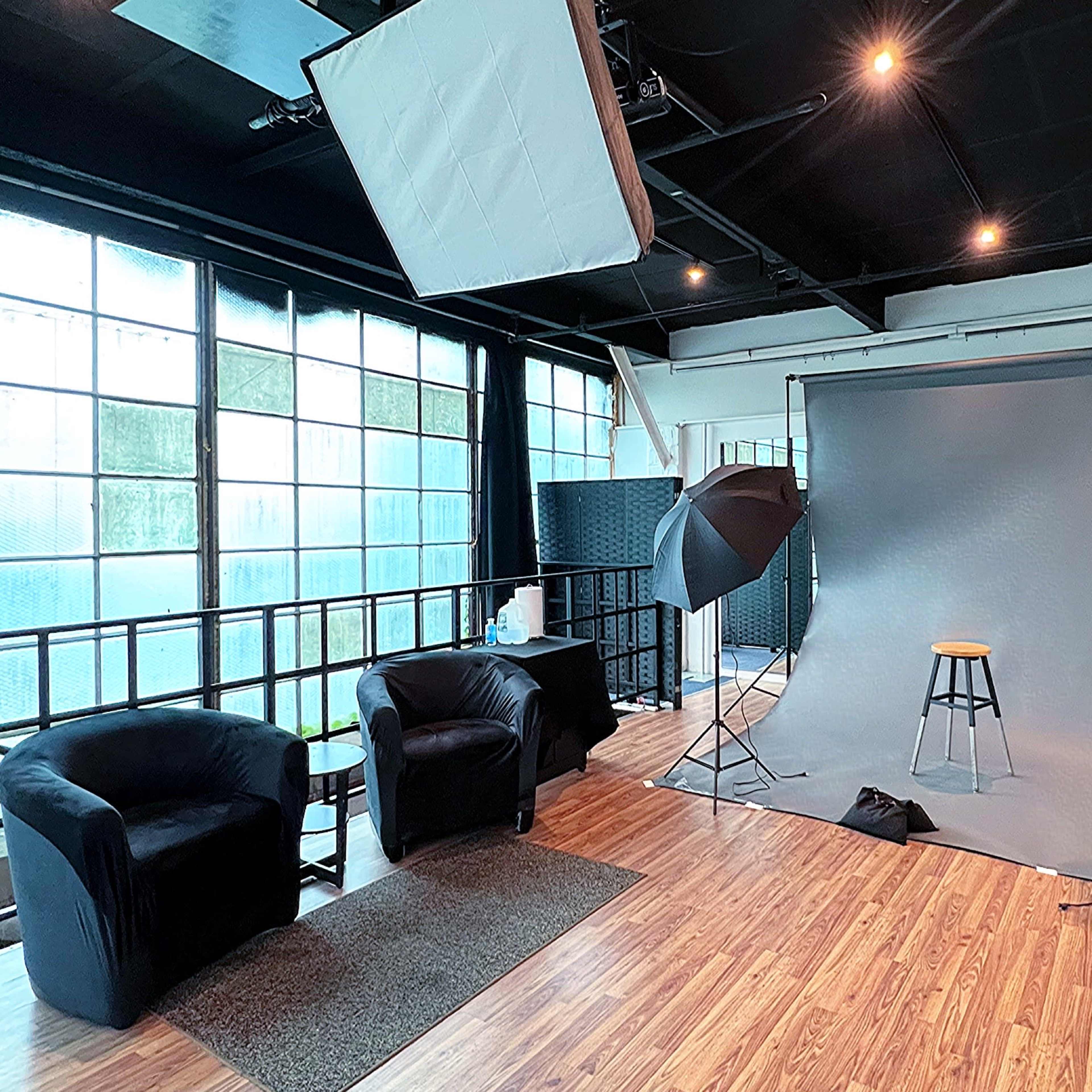 A photography studio with a grey backdrop, two lighting setups, a stool, a black couch, and large windows allowing natural light.