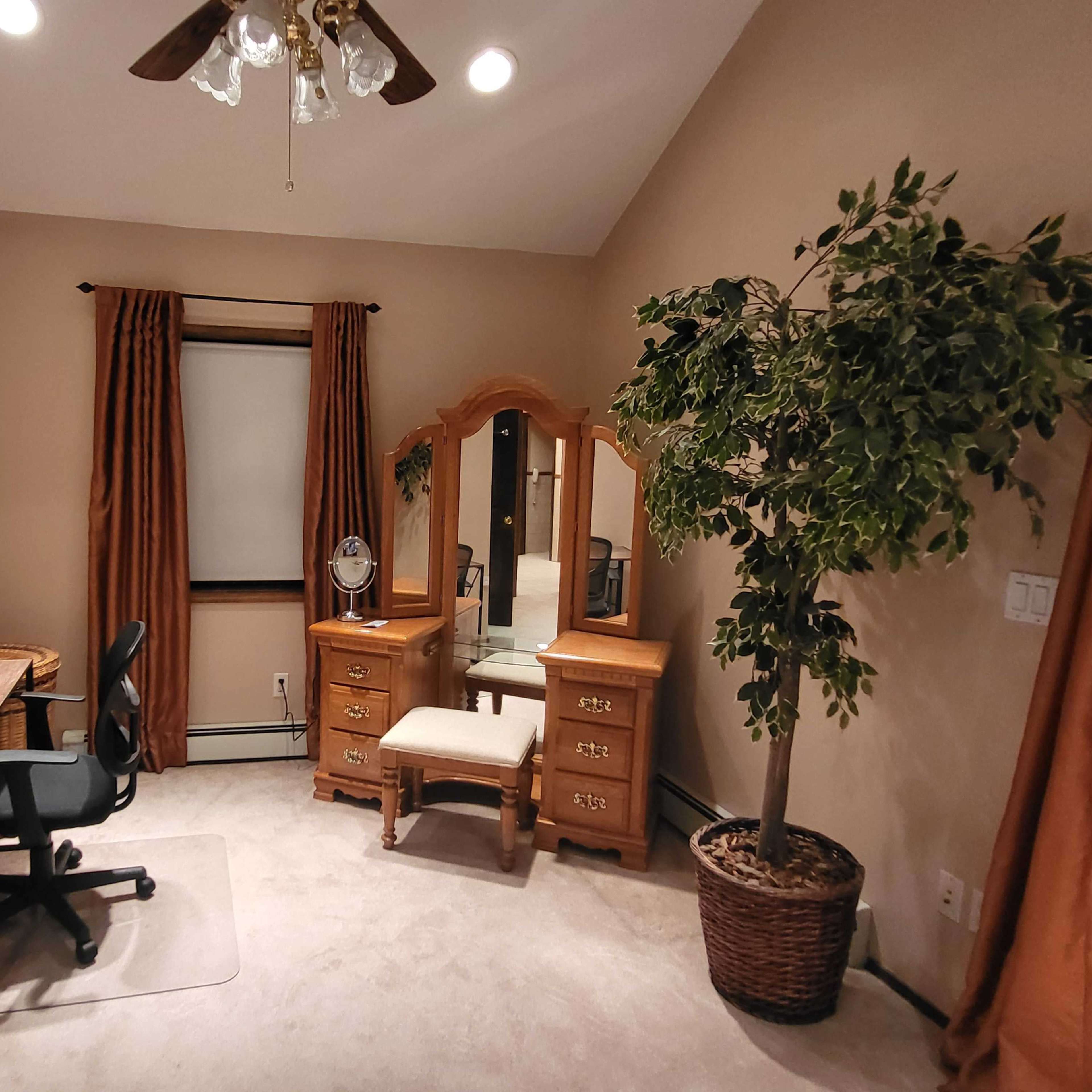 A neatly arranged bedroom with a wooden dresser, a chair and desk, a large window, and a potted plant in the corner.
